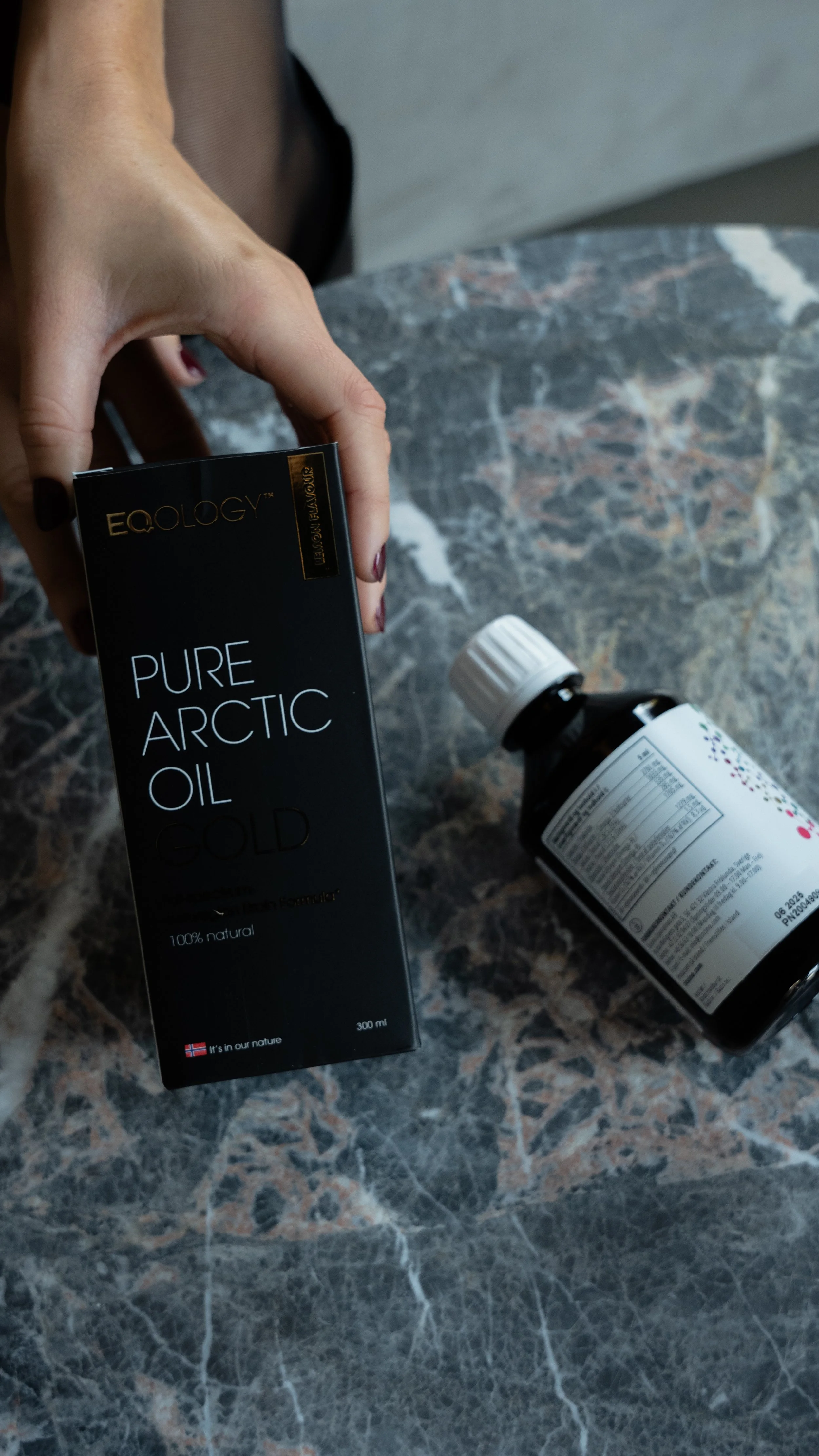 Person holder en sort æske med teksten 'Pure Arctic Oil Cold' og en lille flaske med medicin eller kosttilskud på en marmorbord.
