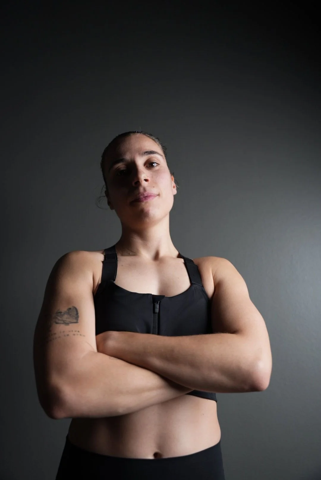 En ung kvinde med arm tatoveringer, iført sort sportstøj, står med foldede arme mod en neutral grå væg.