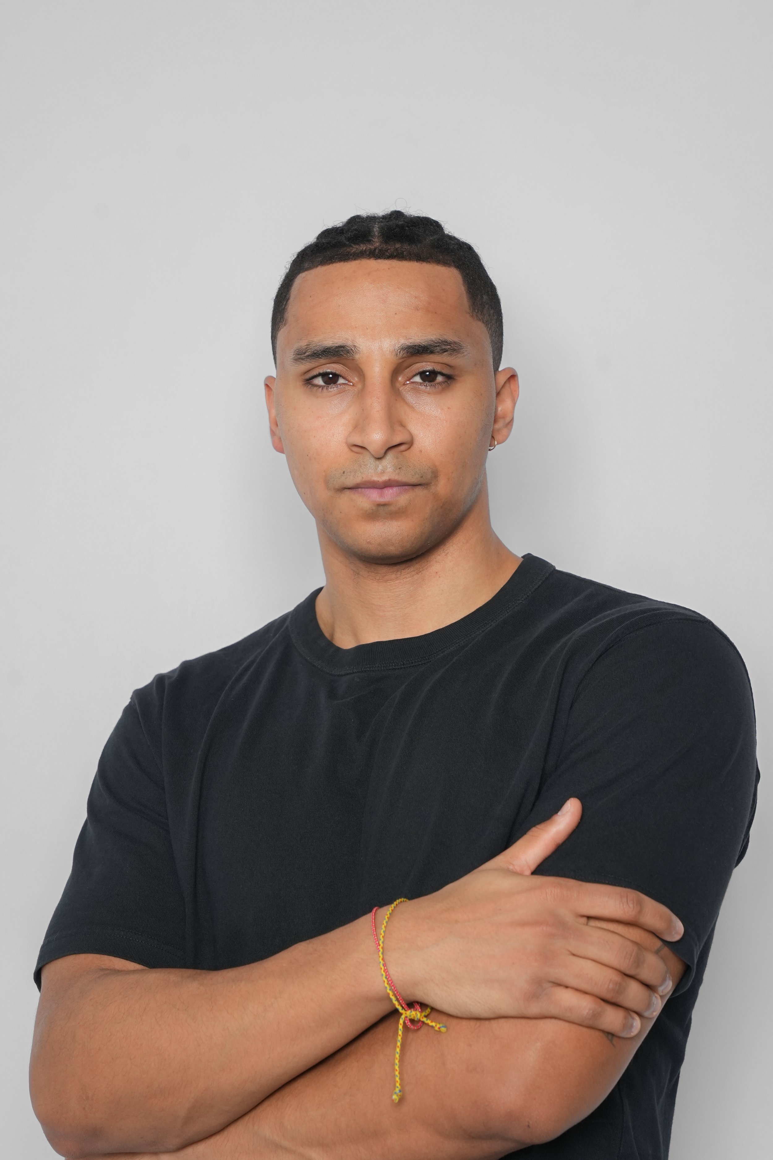 Ung mand med kort, krøllet hår, let skæg, iført sort t-shirt og armbånd, står med armene over kors mod en lys baggrund.