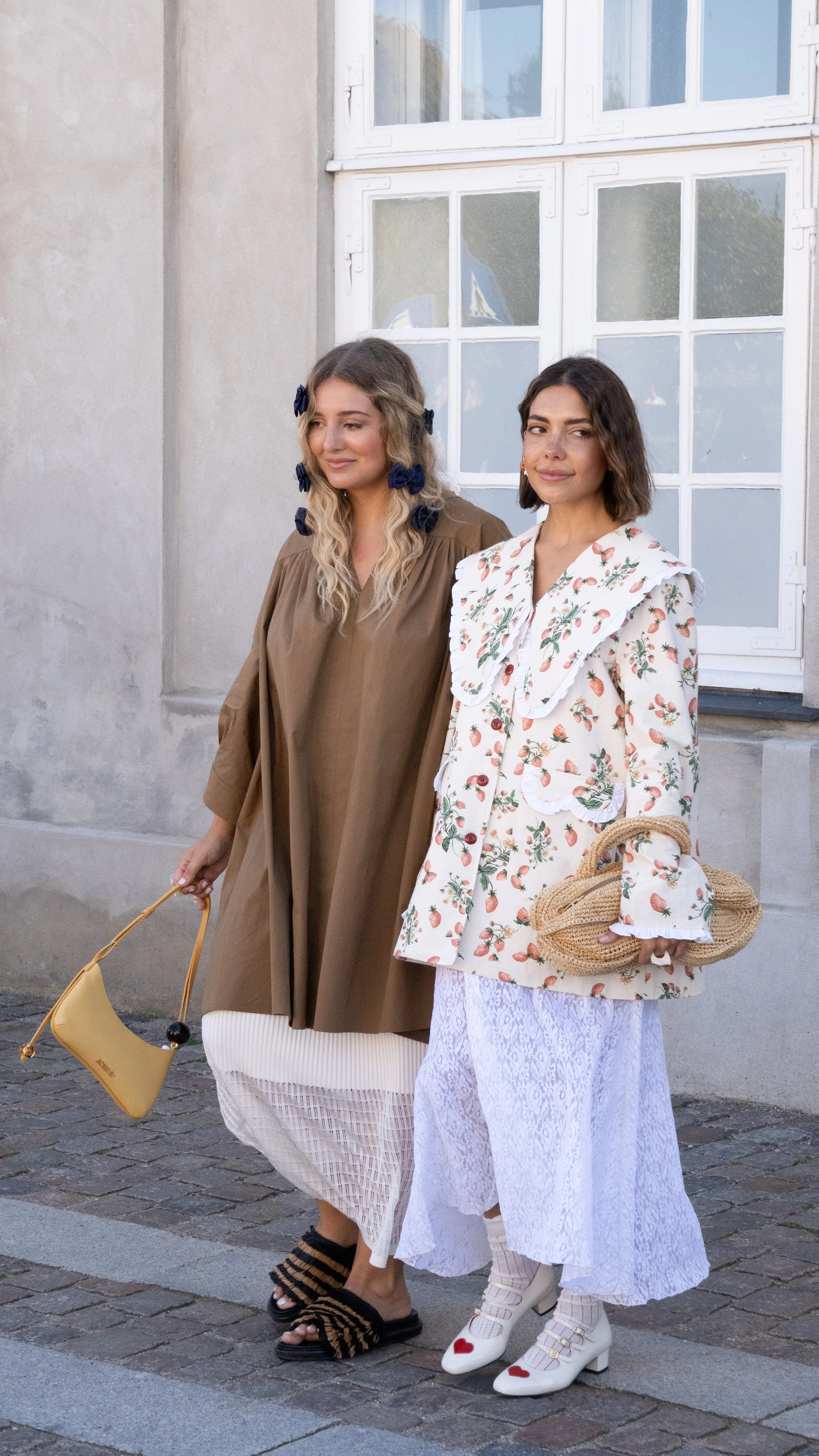 To kvinder går på gaden iført moderigtige outfits med farverige accessories, i en by omgivet af bygninger.