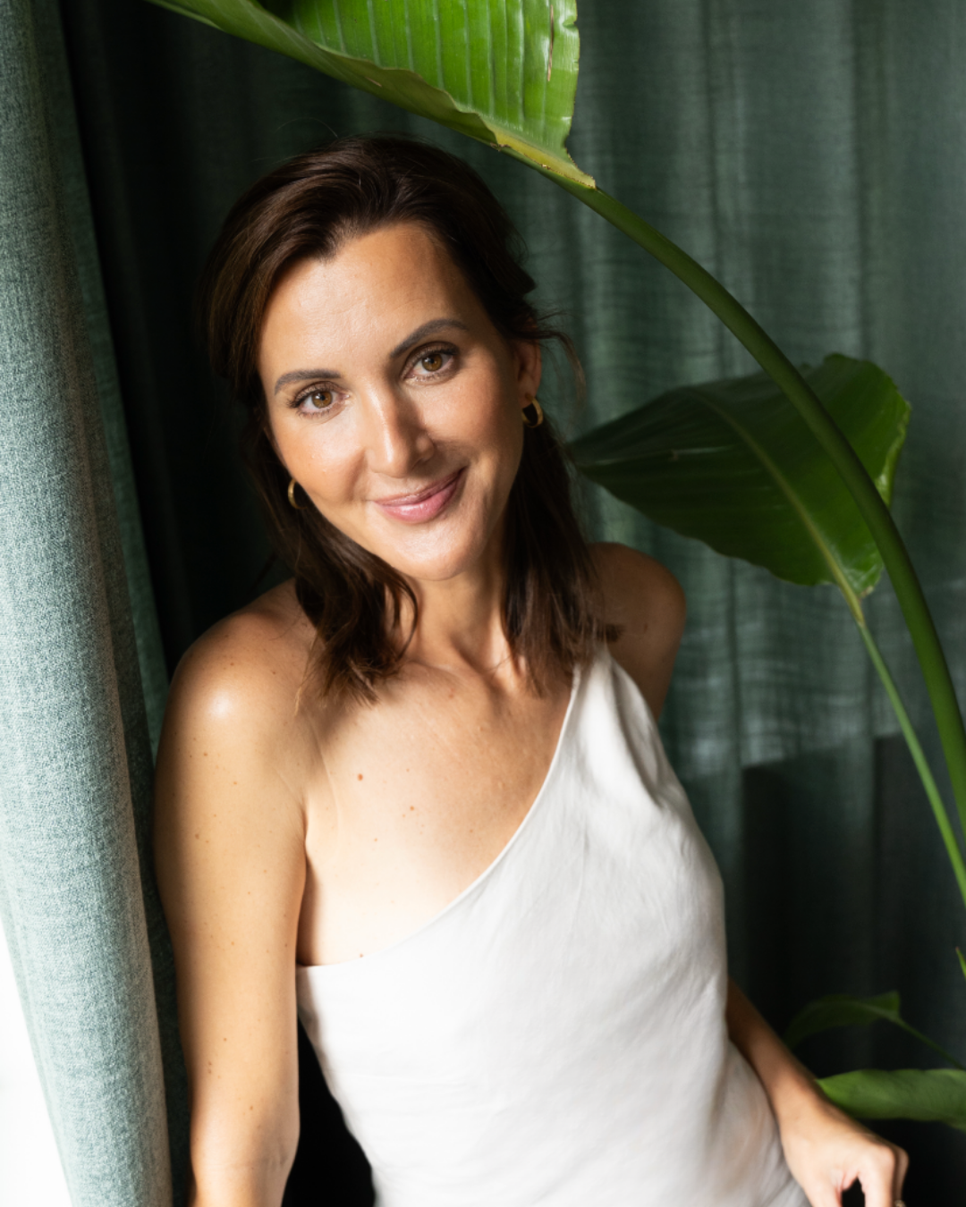 Kvindelig person med brunt, skulderlangt hår, smilende, iført hvid top, omgivet af grønne planter og gardiner.