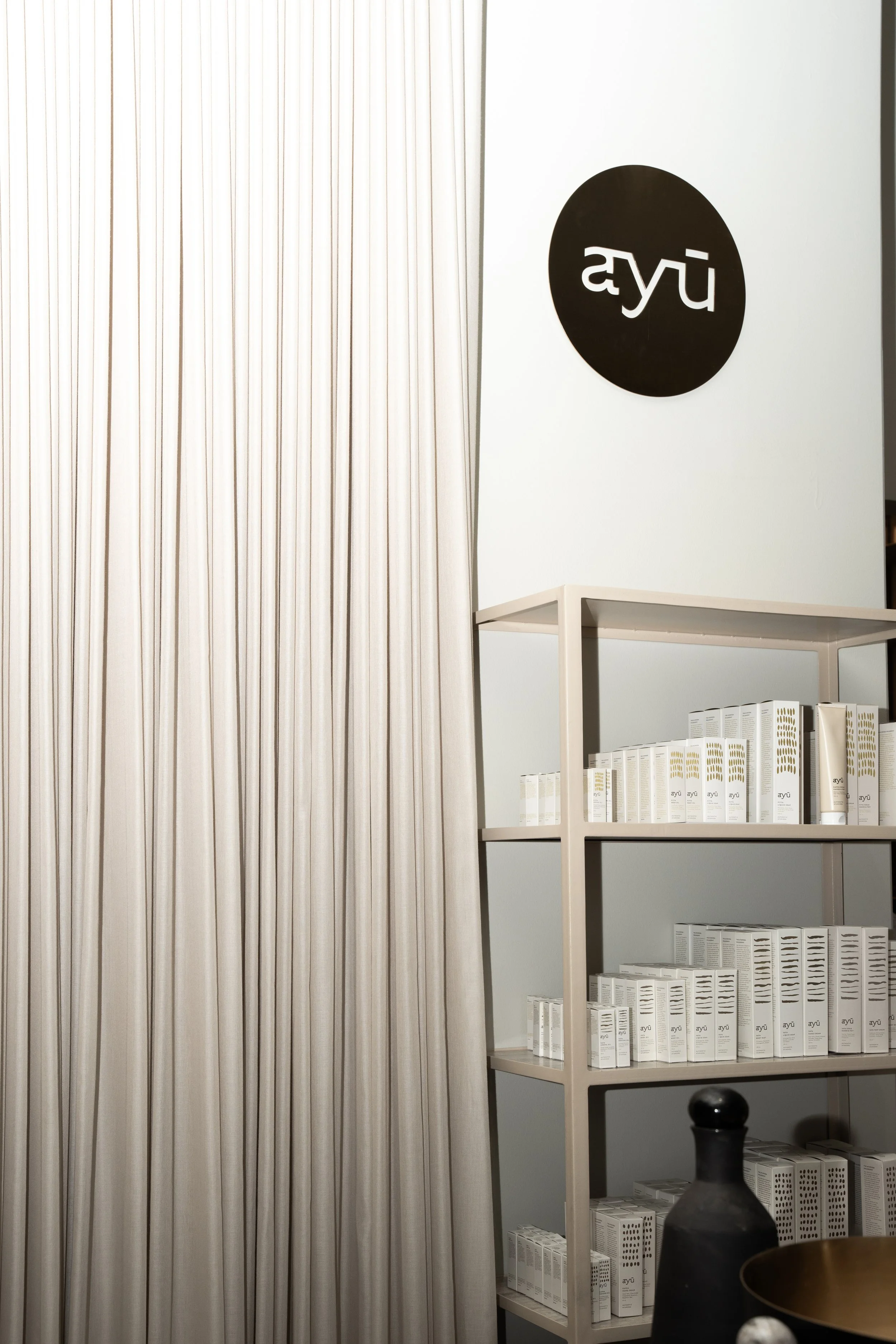 En hvid væg med et rundt sort logo med hvid tekst 'ayu'. Der er en hylde med hvide emballager og en sort dekorativ genstand foran.