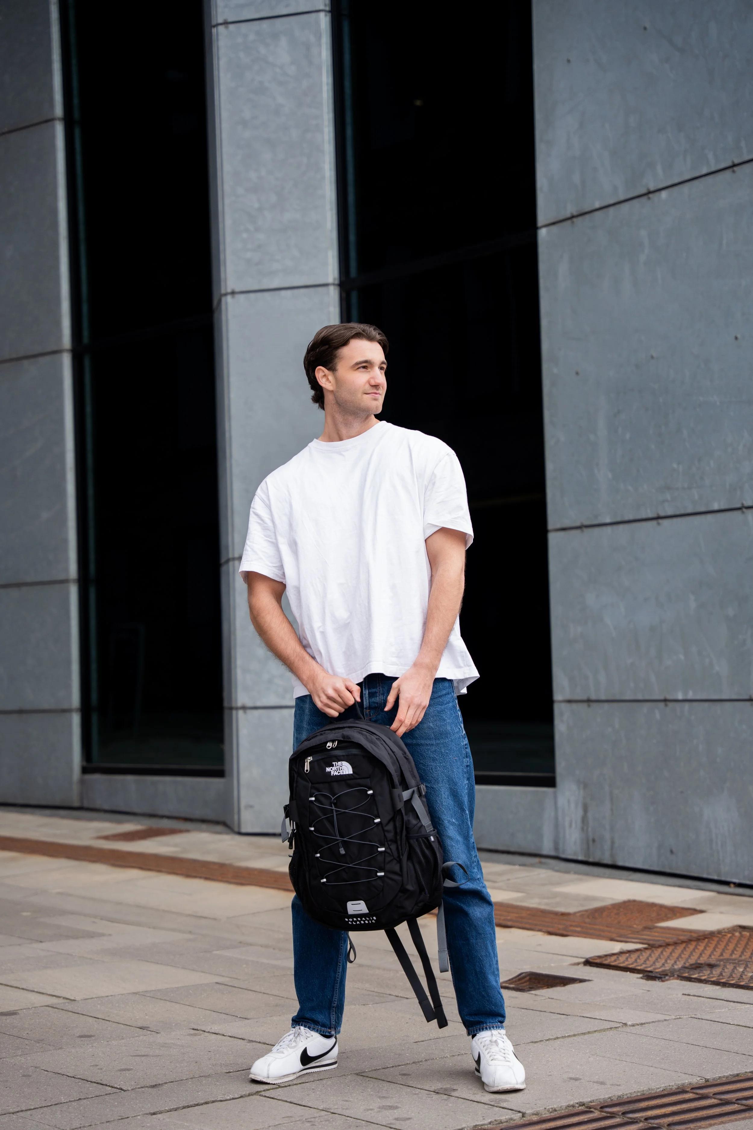 Ung mand i hvid T-shirt og blå jeans står udenfor en moderne bygning, holder en sort backpack.