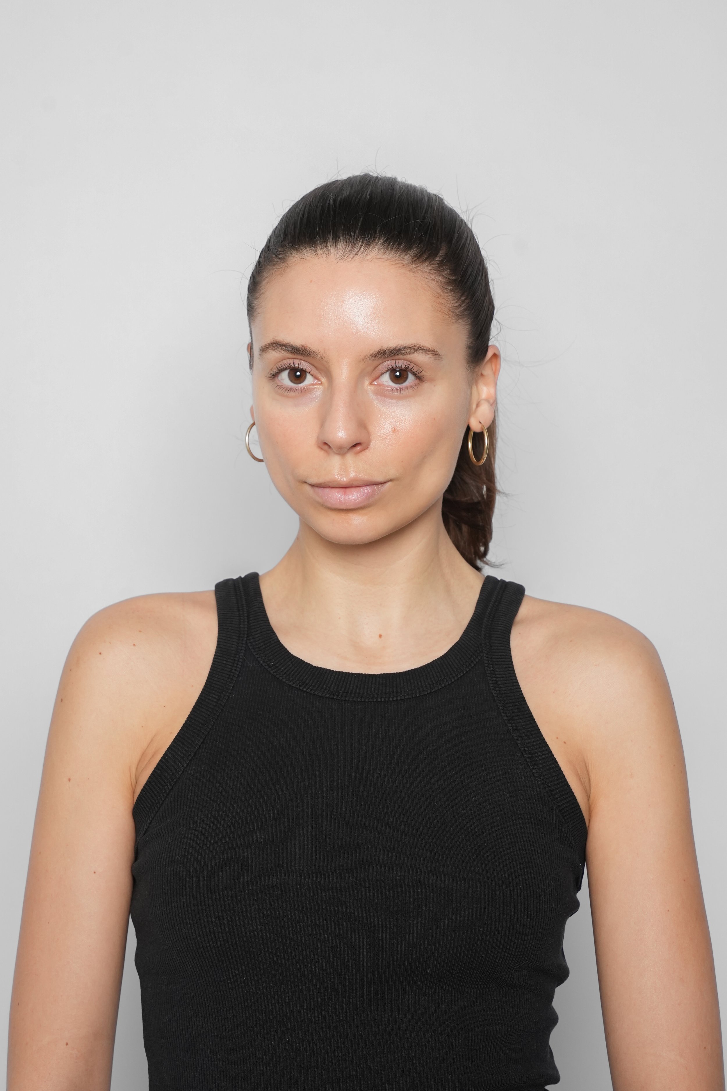 En ung kvinde med mørkt, langt hår i en hestehale, iført sort tanktop og ørestikker, står foran en grå baggrund.