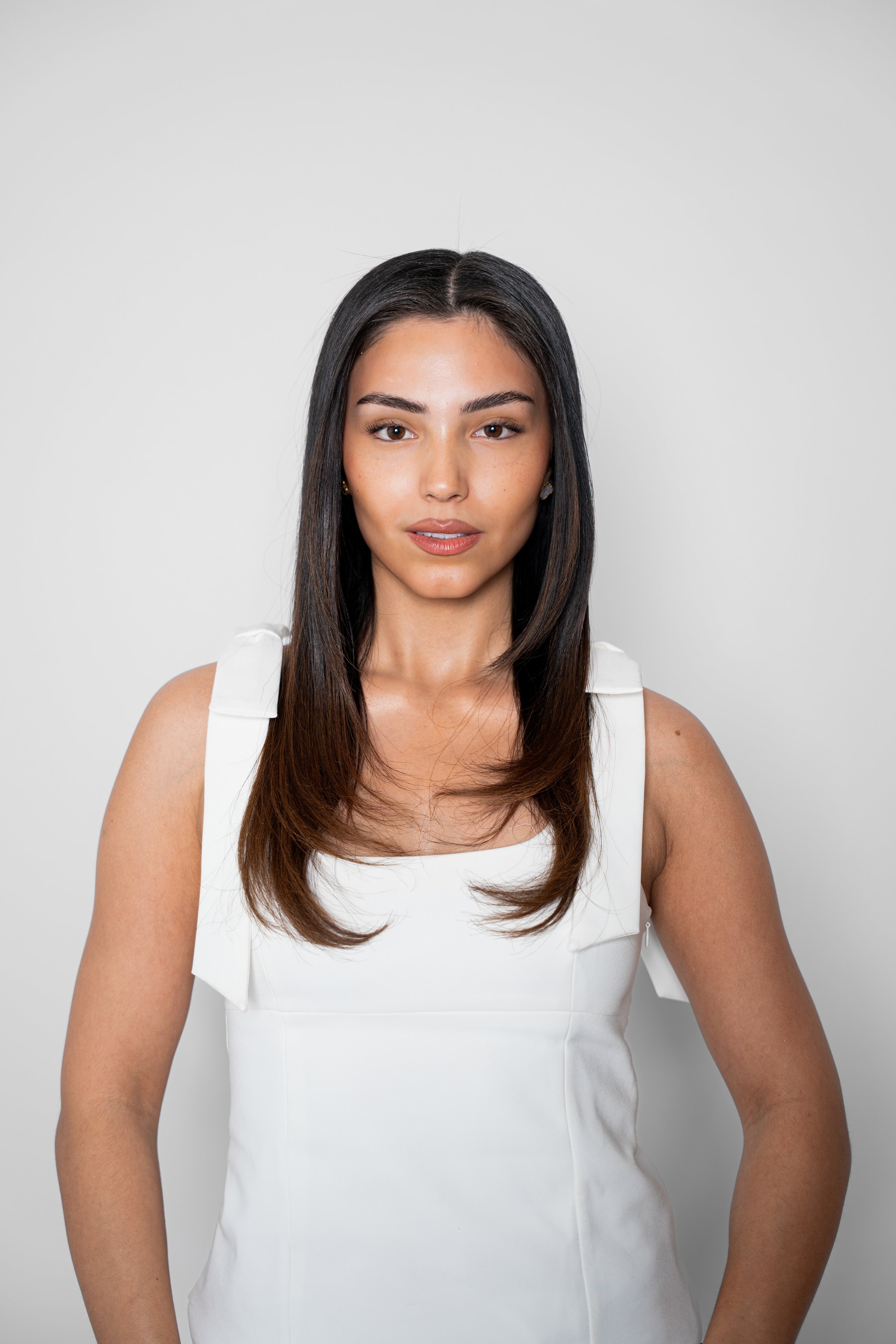 Portræt af en ung kvinde med langt brunt hår, iført hvid kjole, står foran en simpel lysgrå baggrund.
