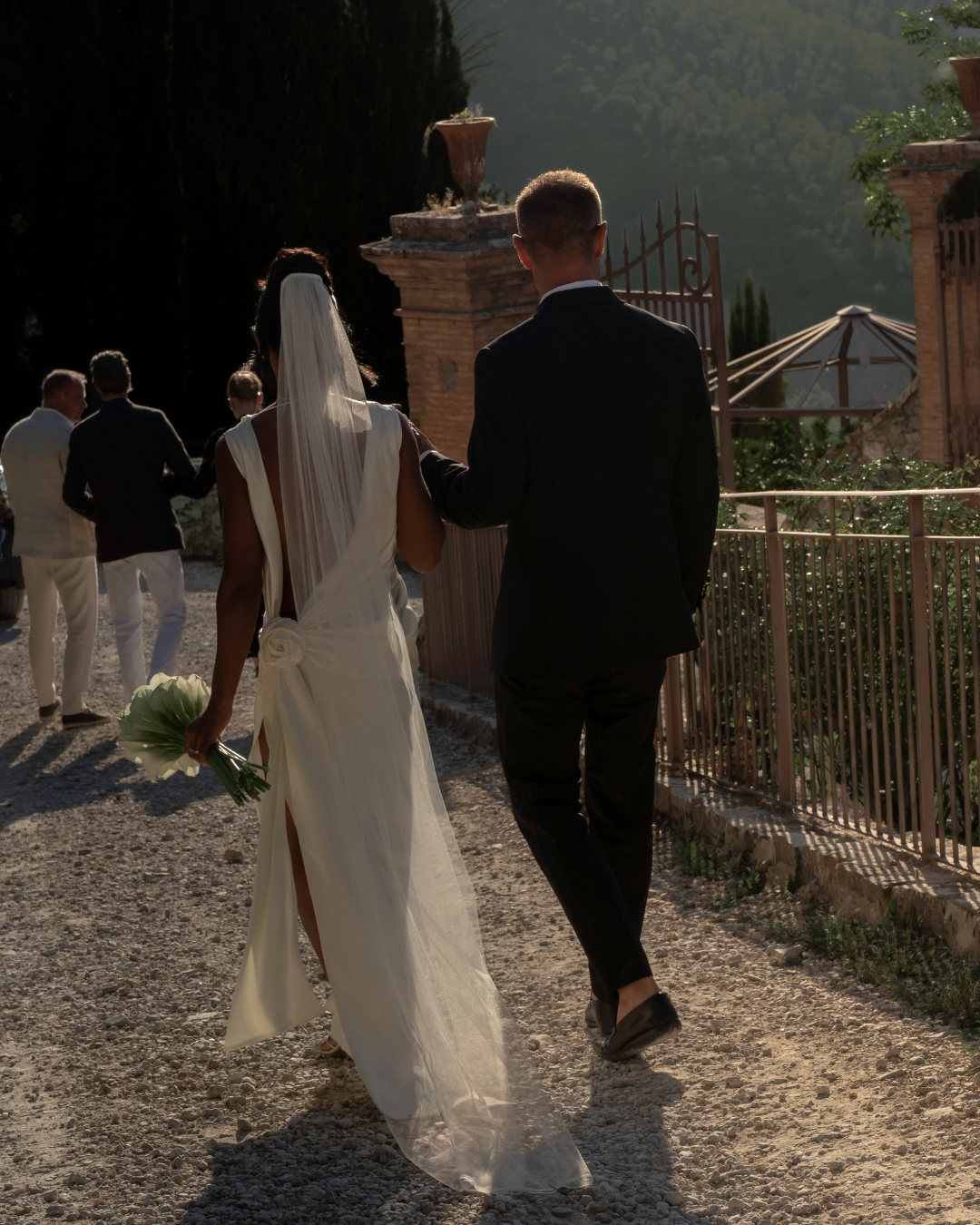 En brud og brudgom går hånd i hånd udendørs, omgivet af andre mennesker, med en historisk mur og grønne træer i baggrunden.