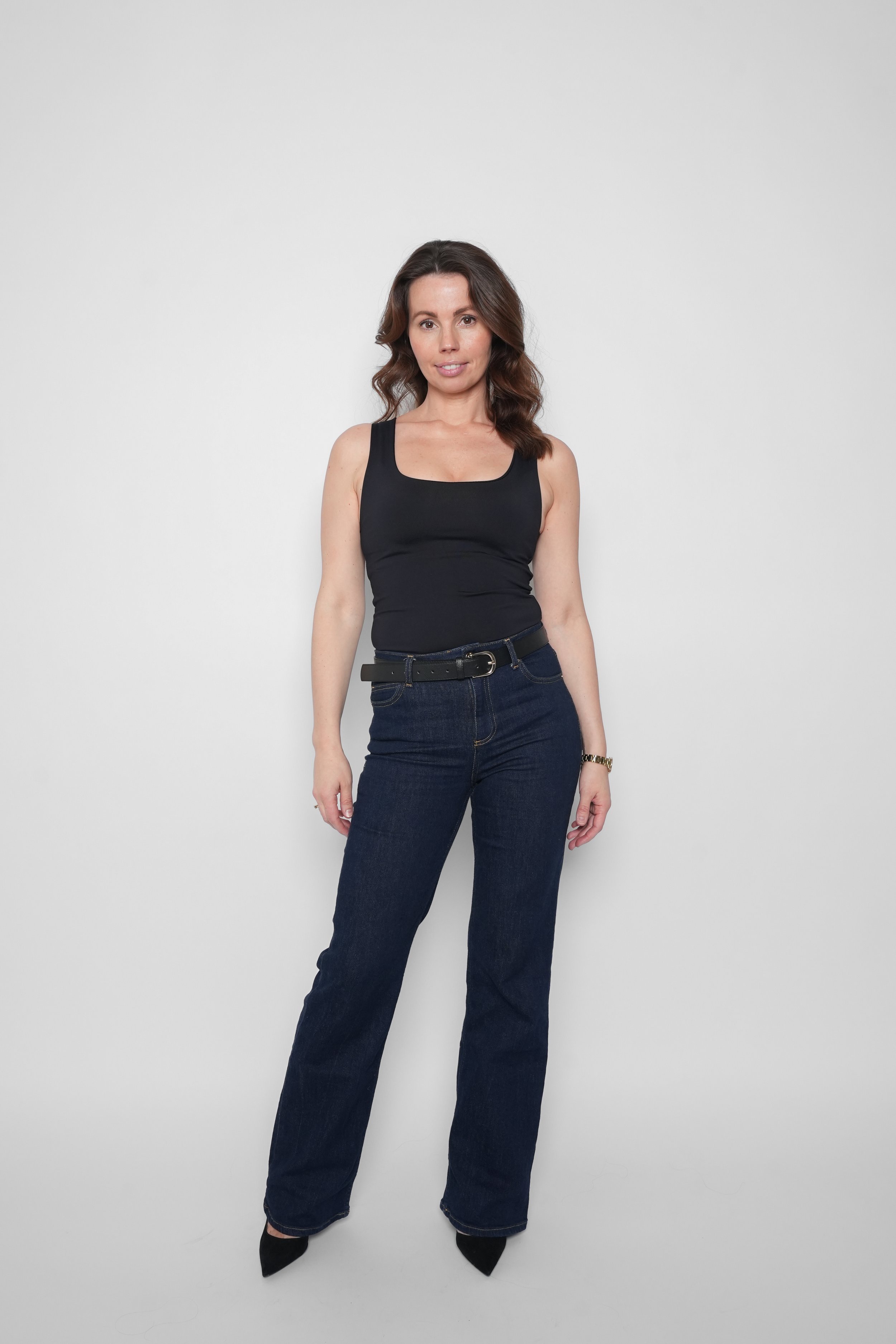 Kvindelig person med brunt, mellemlangt hår iført sort top, blå jeans og sorte klipklapper, står foran en hvid baggrund.