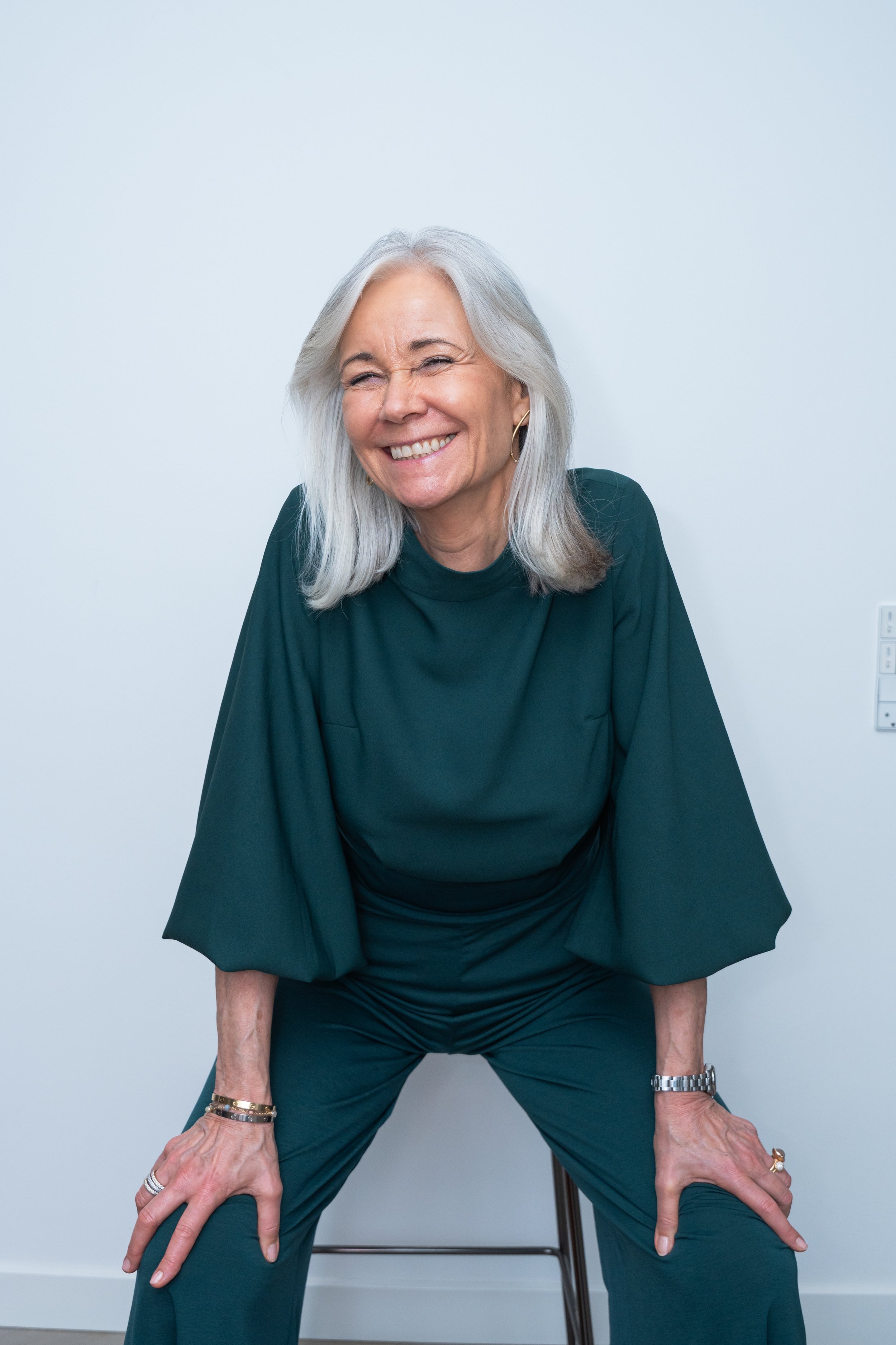 Smilende ældre kvinde med gråt hår i grøn outfit, siddende på en stol mod en hvid væg.
