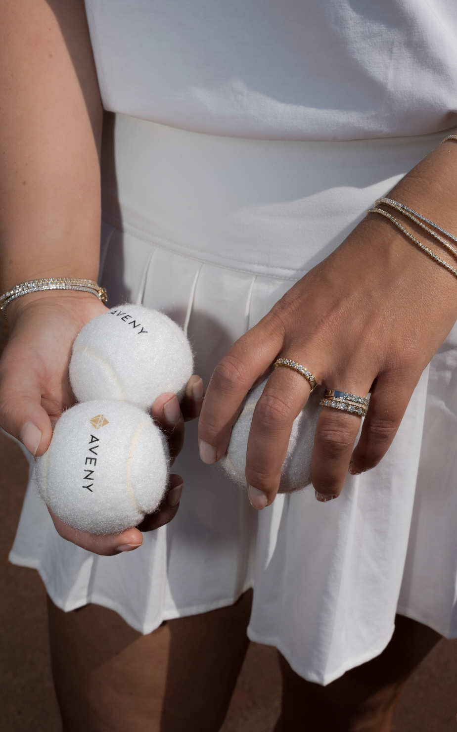 En person i hvidt tøj holder to tennisbolde med logoet 'AVENY', mens de på den anden hånd har tre ringer og et tynd armbånd.
