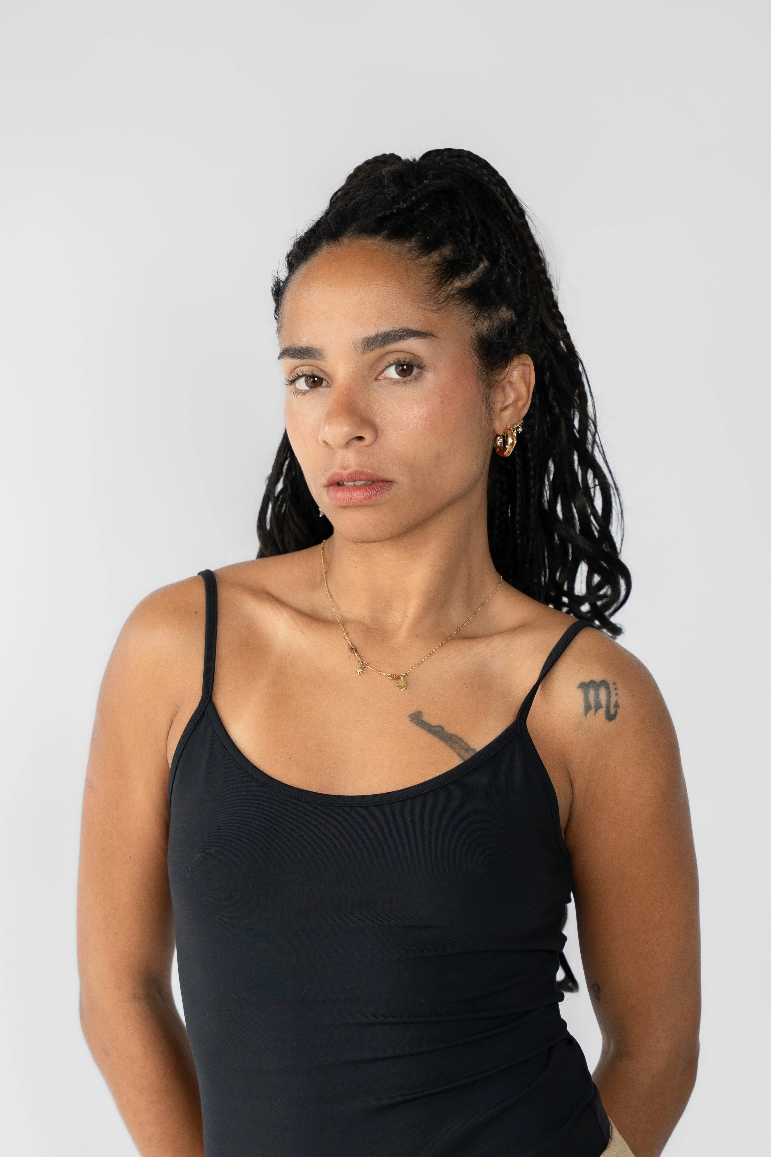 Portræt af en kvinde med dreadlocks iført sort top, guldbjælder, og halskæde, stående mod en hvid baggrund.