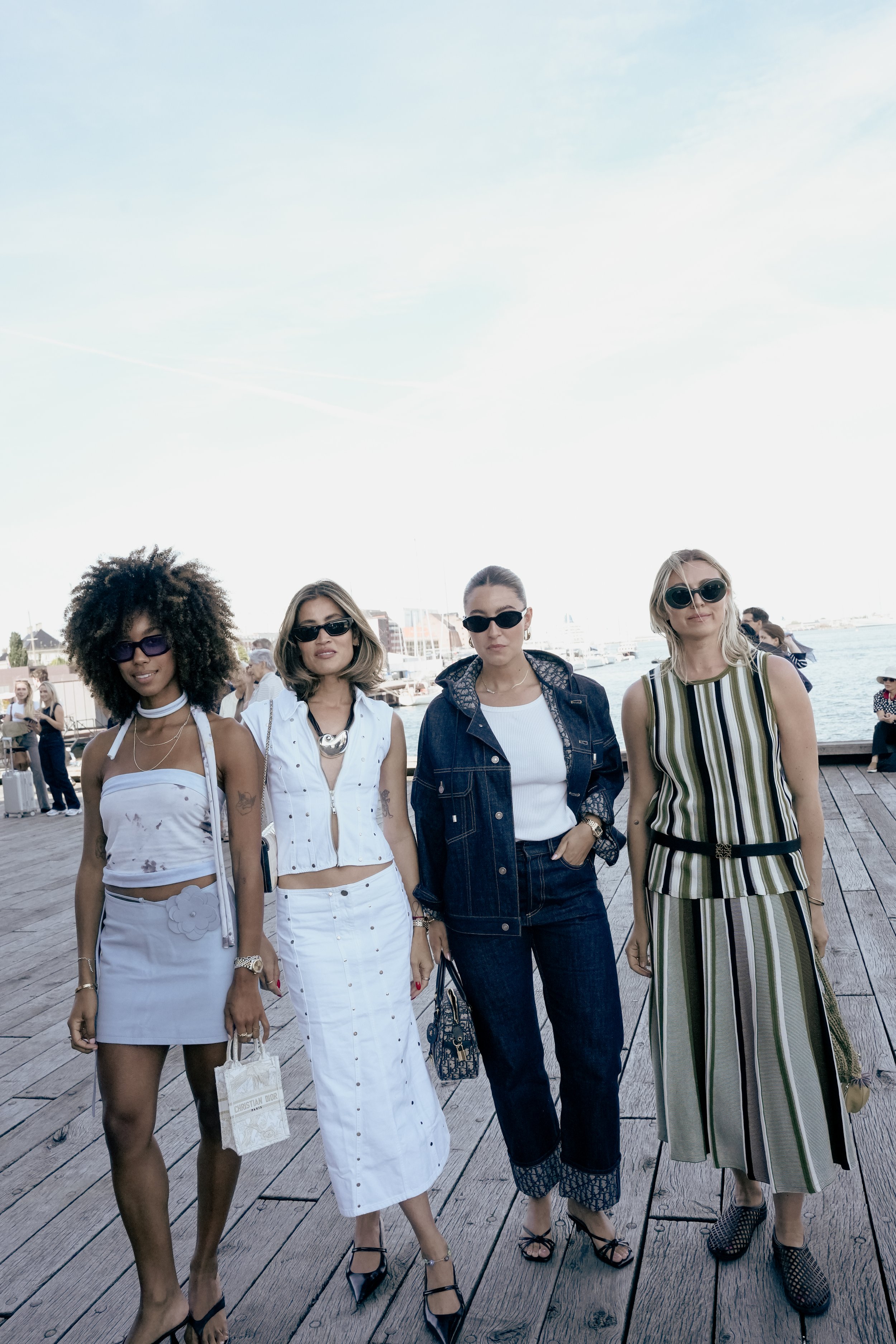 Fire kvinder spaserer på en dokke ved vandet, alle i solbriller og sommeroutfits.