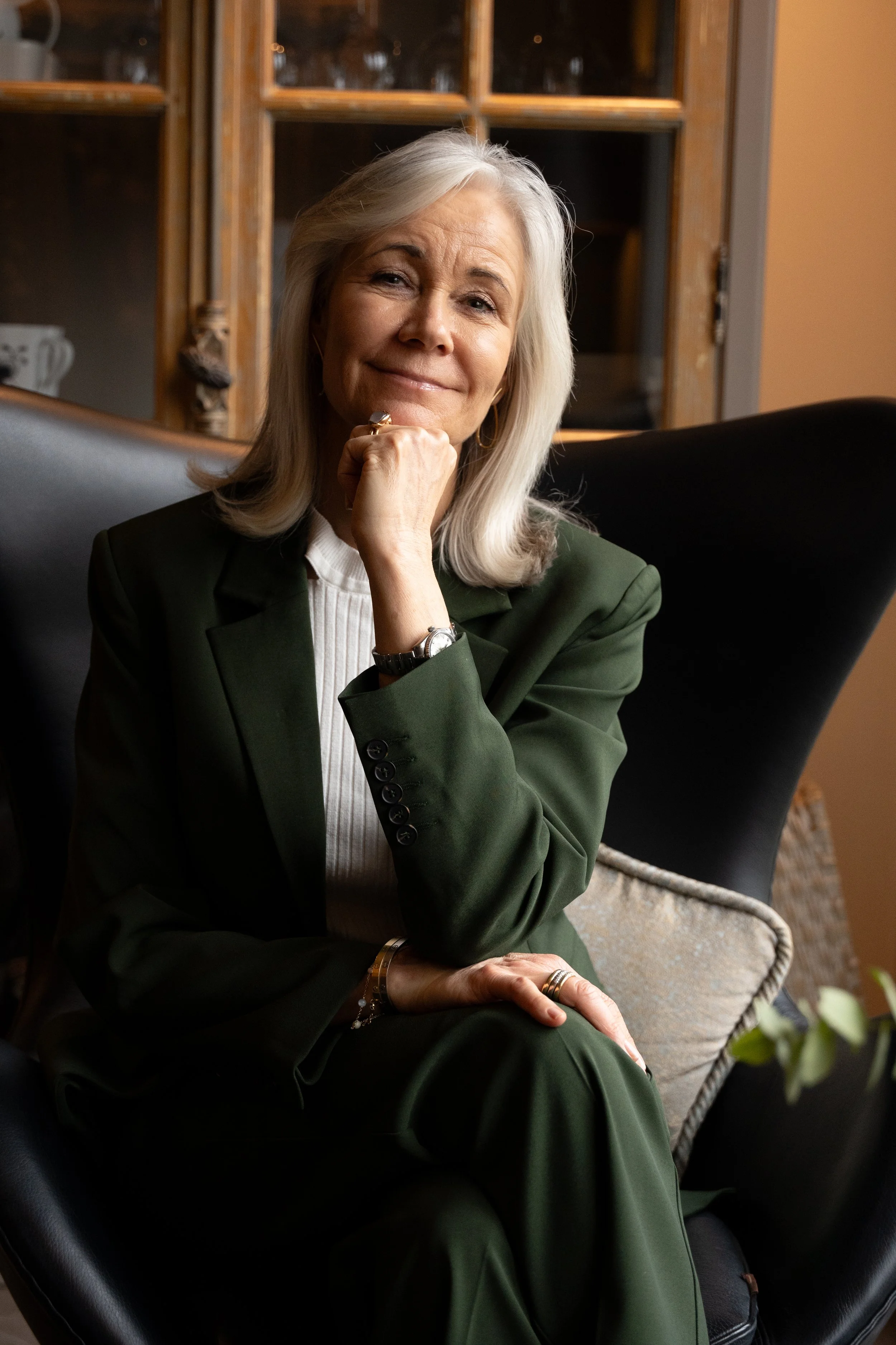 En ældre kvinde med gråt hår, iført en grøn jakkesæt, sidder i en stol med en taknemmelig eller kløgtig udtryk. Bag hende er en skænk med glas og porcelæn.