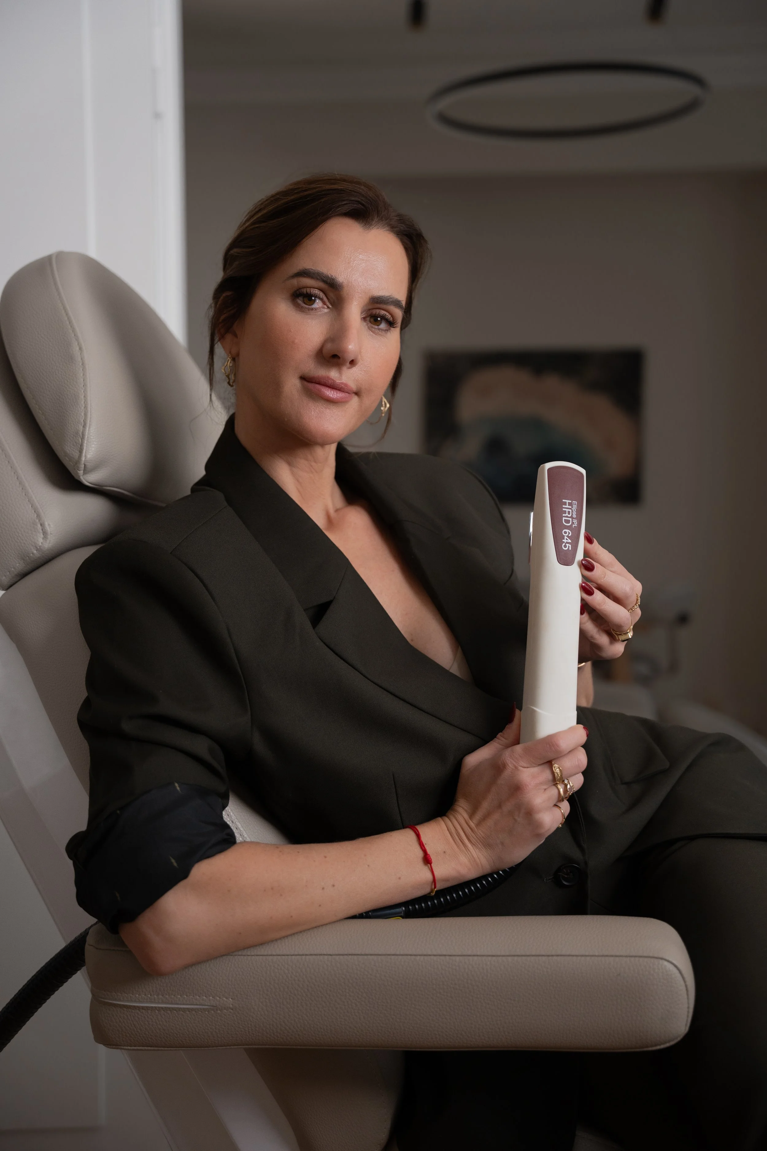 Kvinde sidder i en komfortabel lænestol med en medicinsk enhed i hånden, iført en sort blazer, i et moderne klinik.