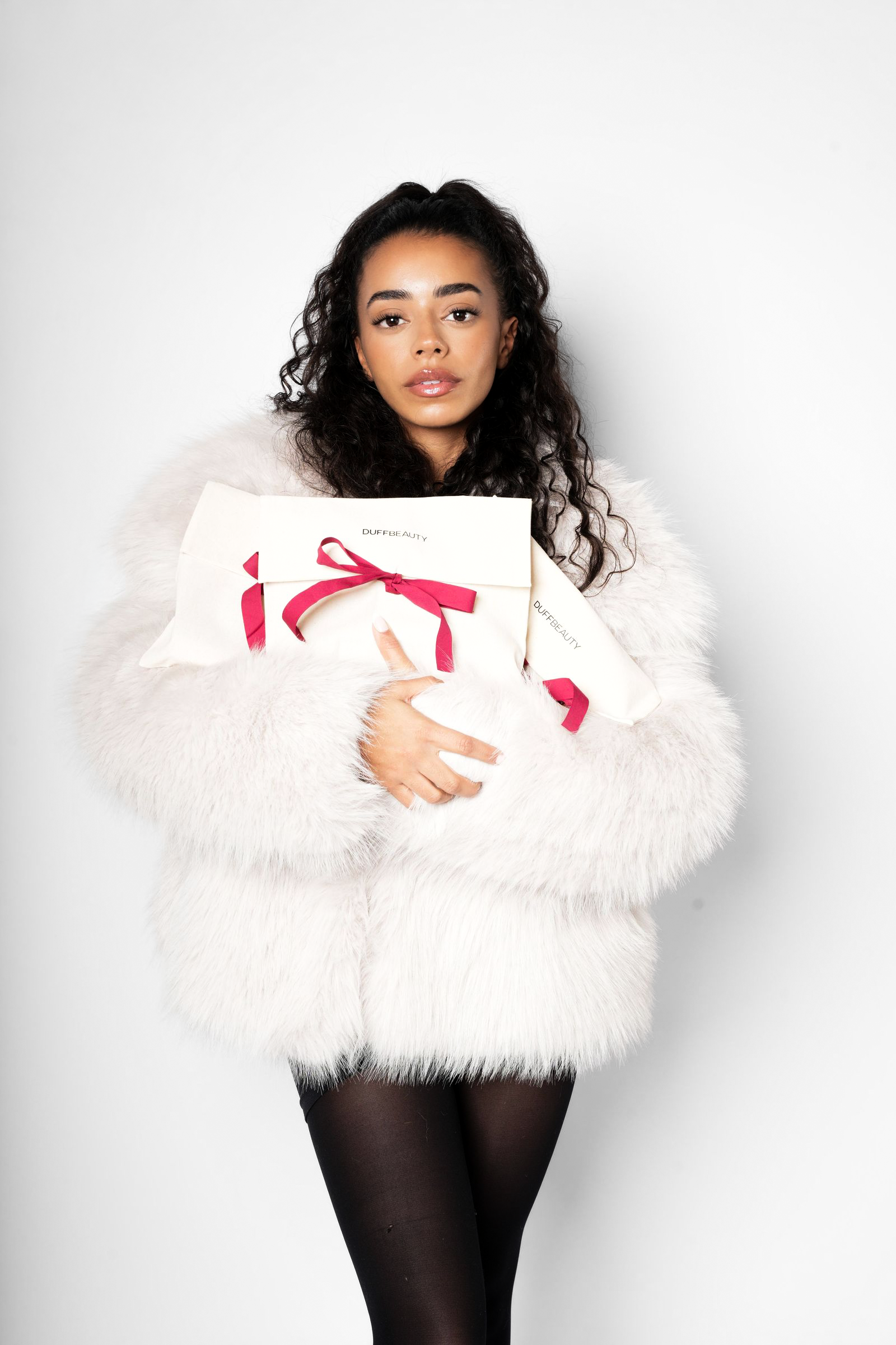 Kvindelig model med mørkt, krøllet hår iført hvid pelsjakke, der holder en gavekasse, mod kamerafront mod en hvid baggrund.