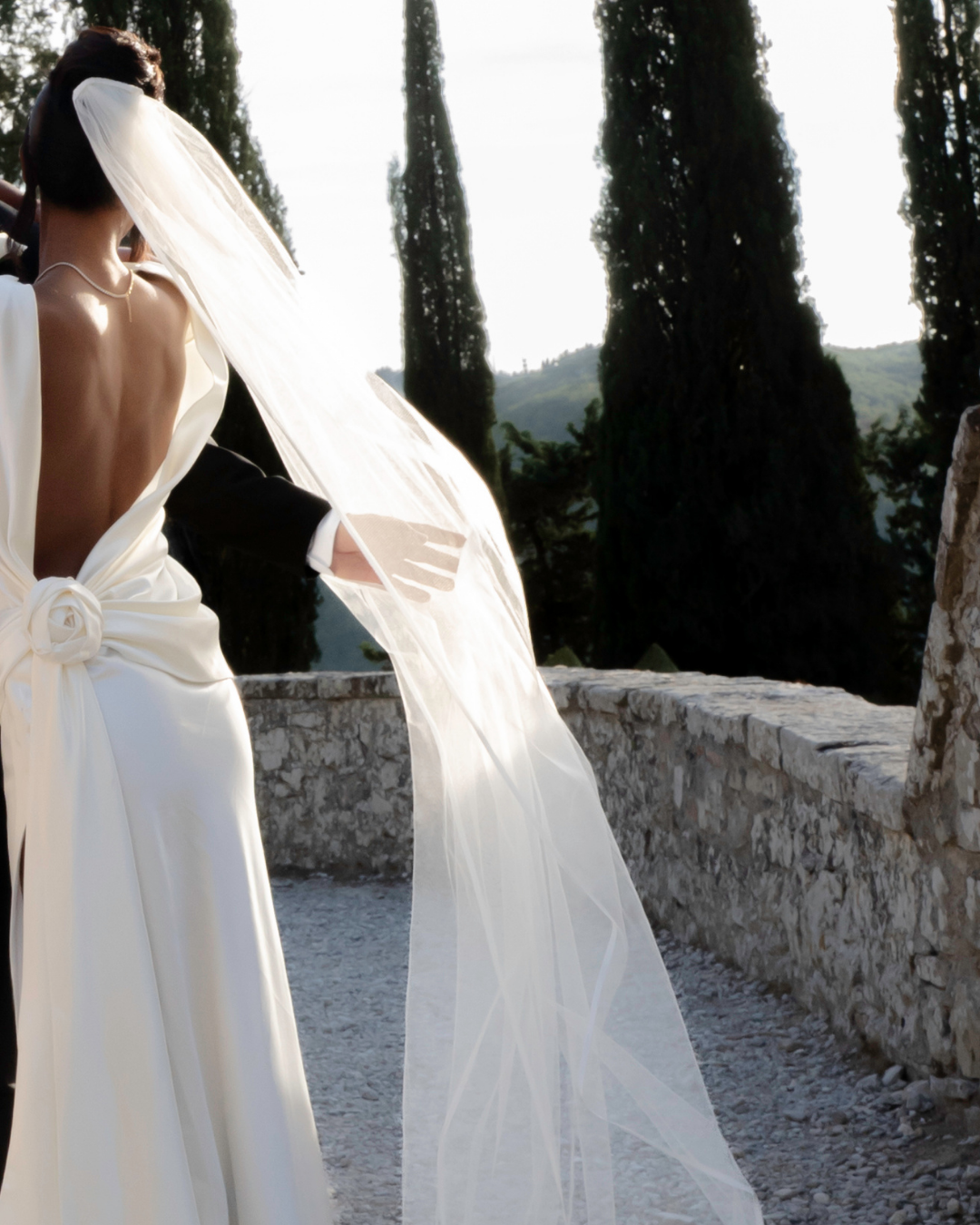 Kvindelig person i brudekjole med veil, taget udendørs ved en mur, med træer i baggrunden.