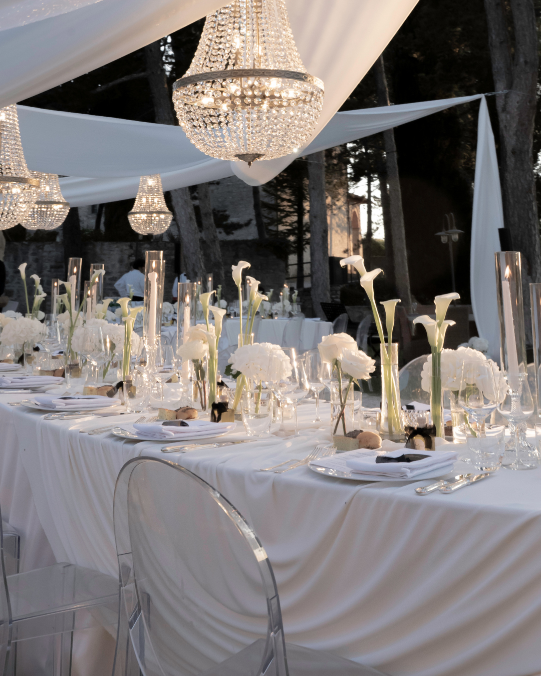 Elegant udendørs middagopdækning med hvid dug, høje hvide blomsterdekorationer, stearinlys og lysekroner, omgivet af træer.