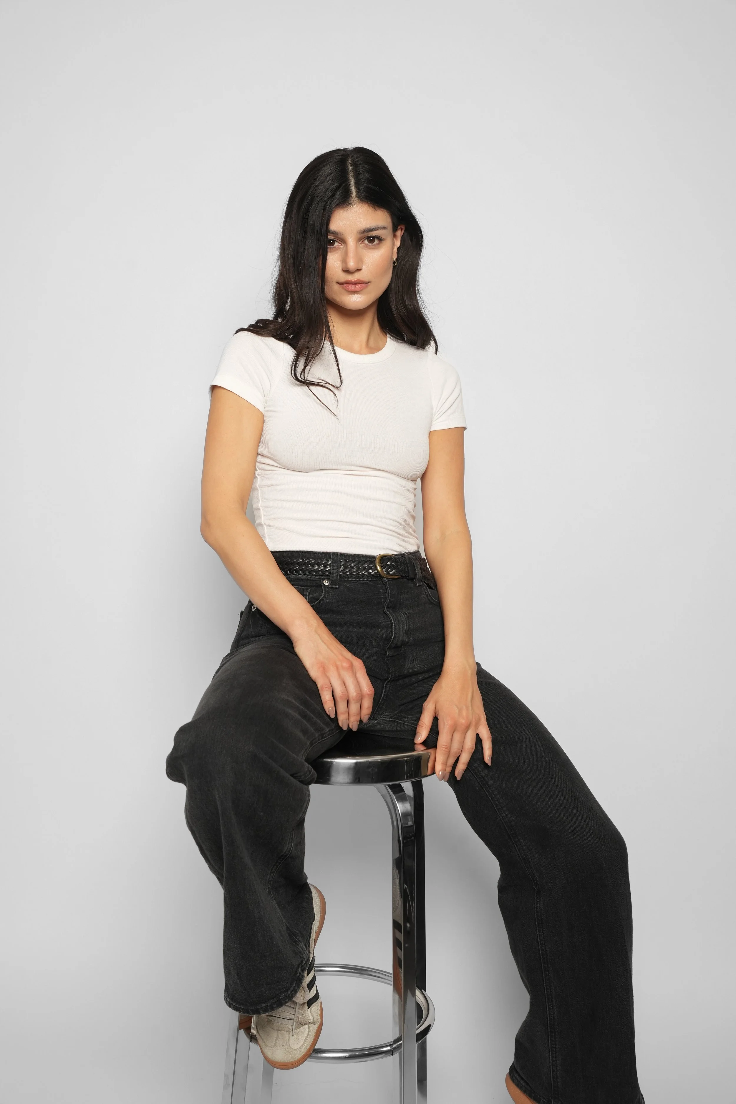 En ung kvinde sidder på en barstol foran en neutral grå baggrund, iført en hvid T-shirt, sorte bukser og beige sneakers.