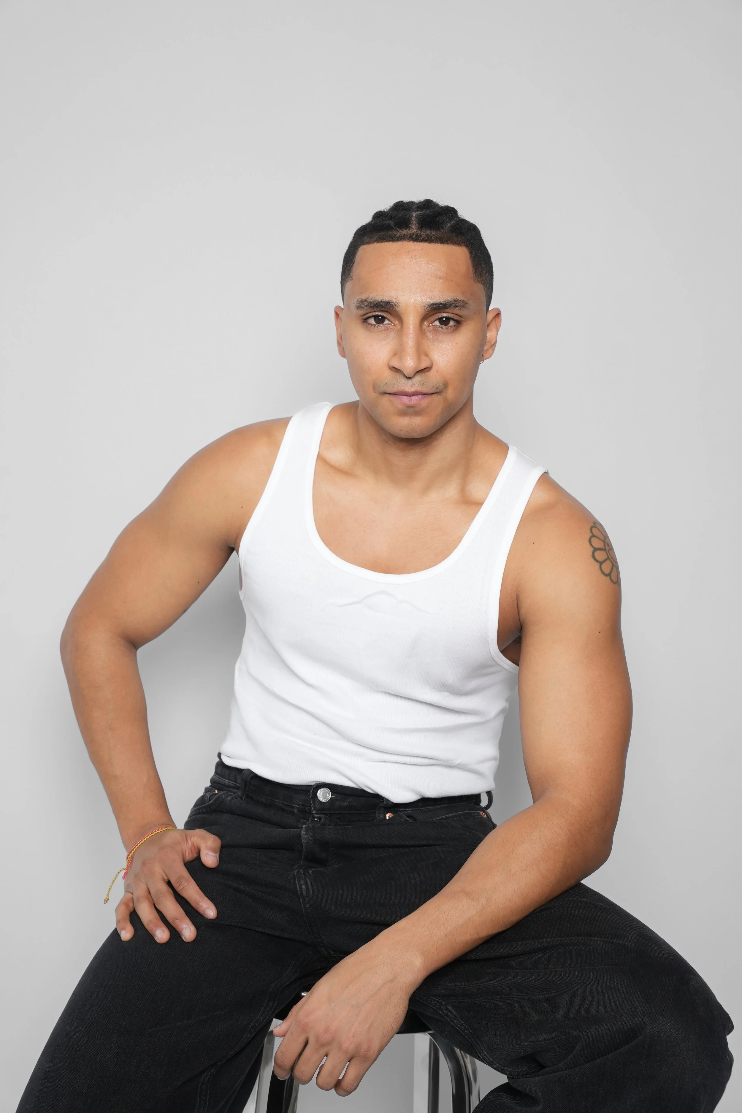 Ung mand i hvid tanktop og sorte bukser sidder på en stol mod en lys grå baggrund.