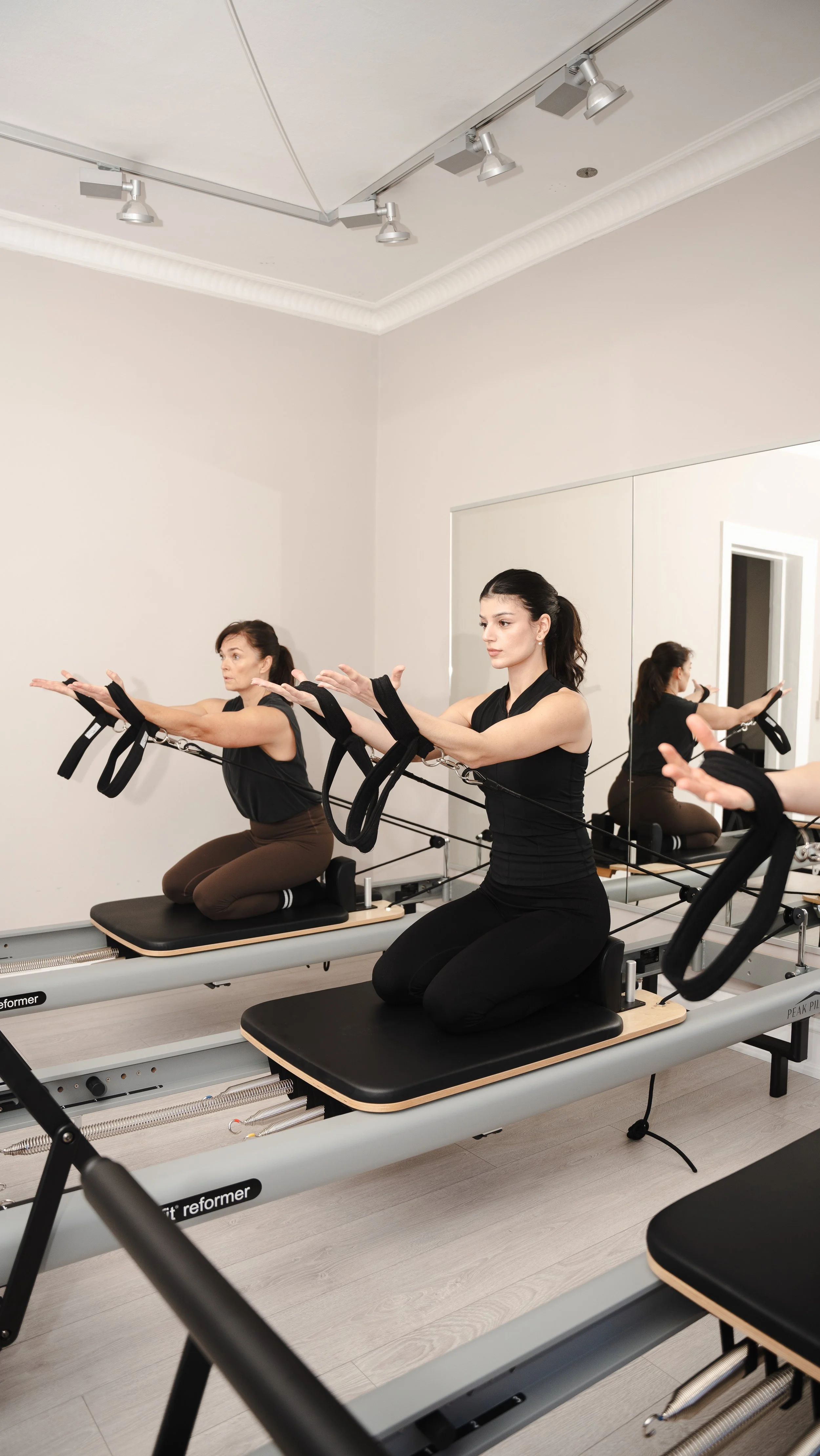 To kvinder træner på reformer i et fitnesscenter, set i et rum med spejlvæg.