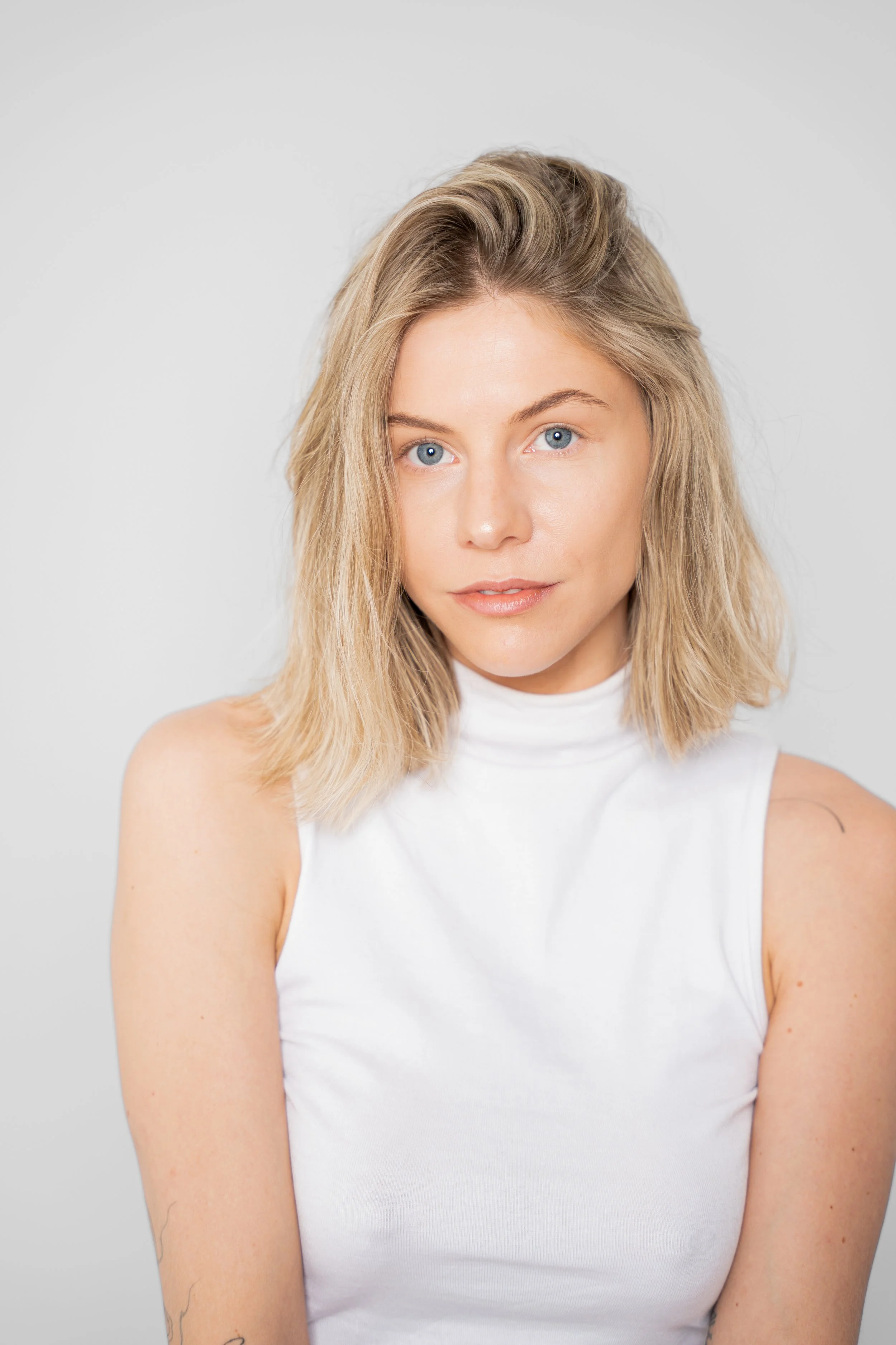 Portræt af en ung kvinde med blond hår og blå øjne iført hvid top mod en neutral baggrund.