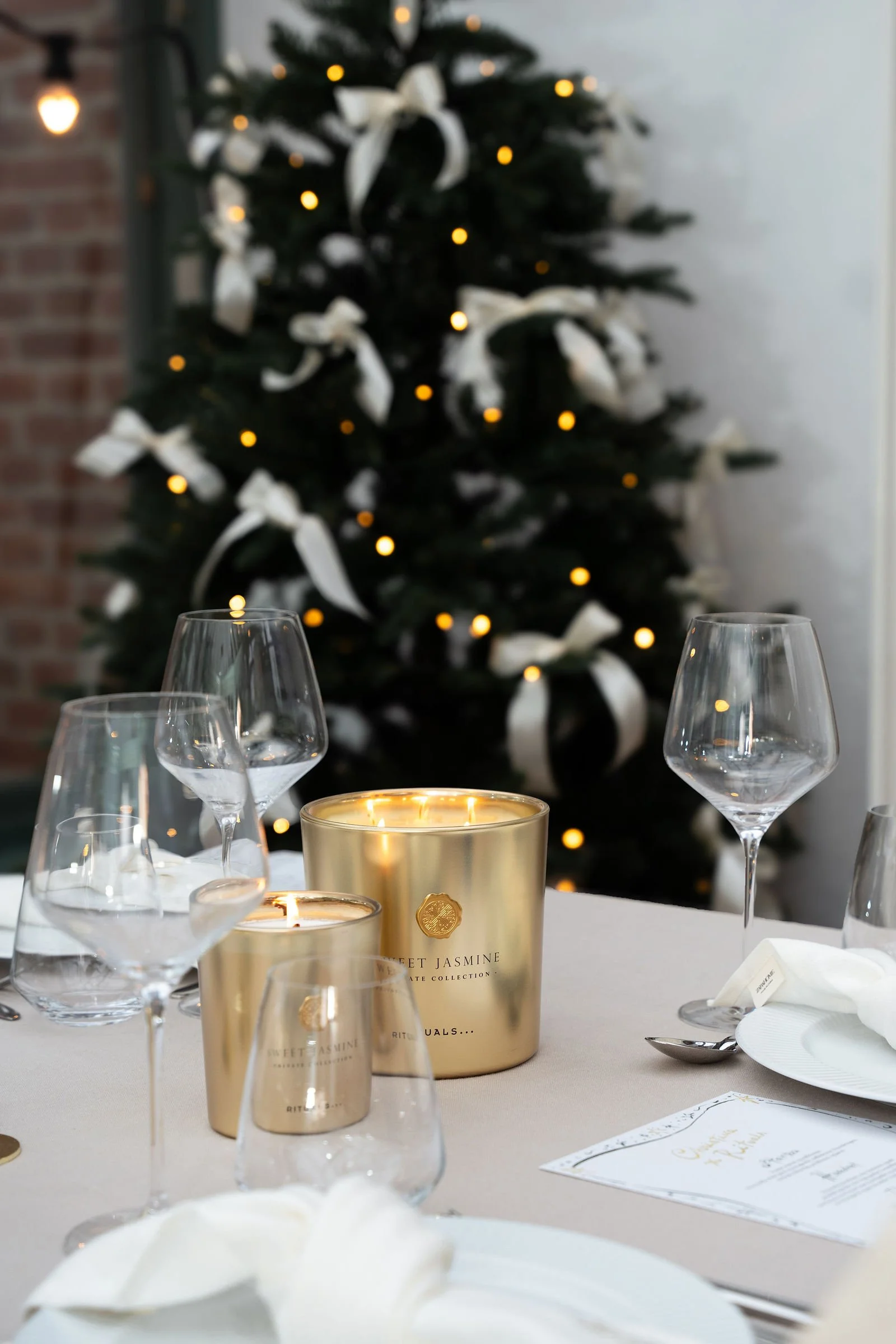 Festligt middagsbord med gyldne lysestager, tomme vinglas, servietter og menu, med juletræ pyntet med hvide bånd og små gule lys i baggrunden.