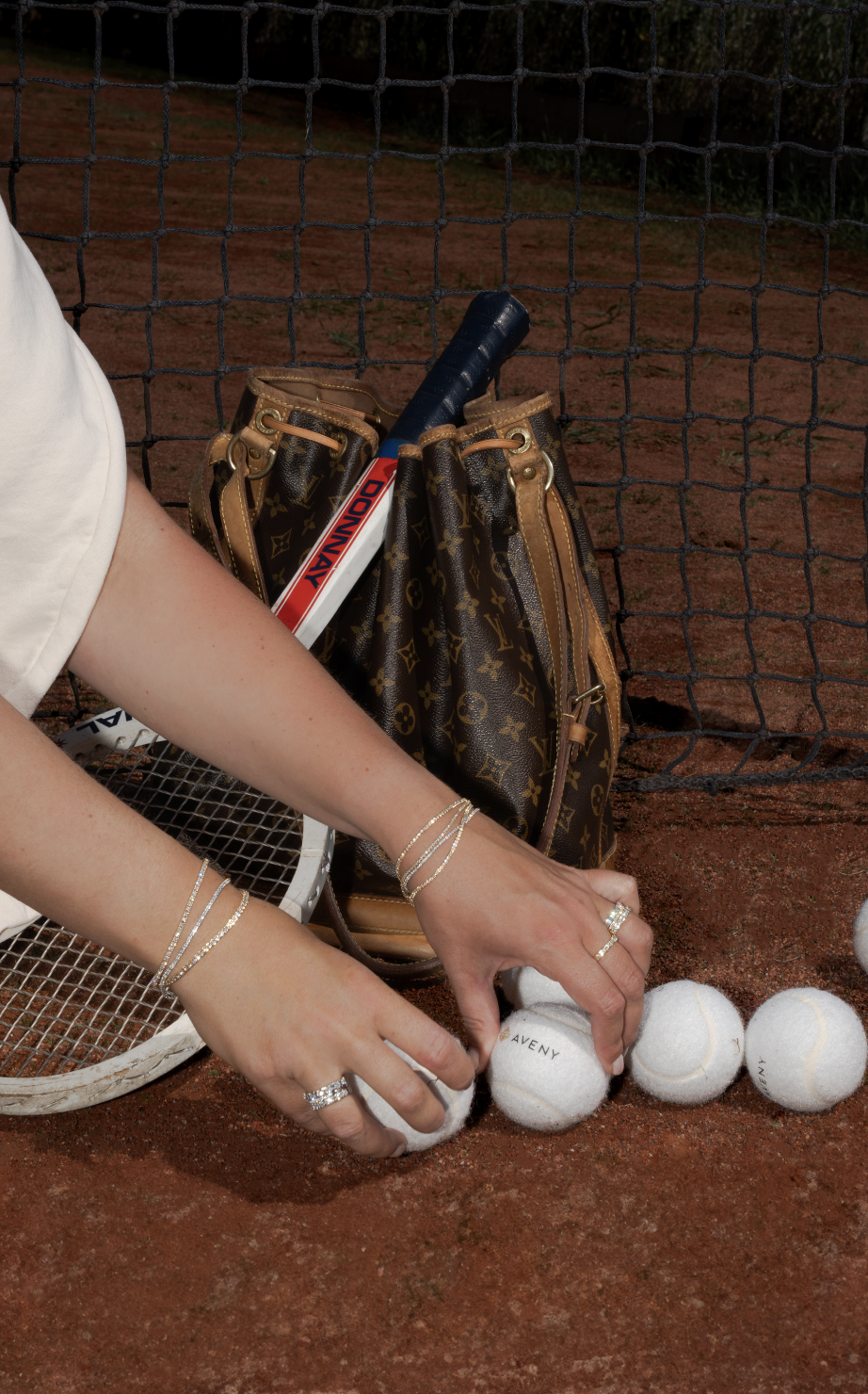 En person placerer tennisbolde på jorden ved at lægge dem fra hinanden. Der er en en taske med louis vuitton-mønster og en tennisbold med 'AVENY' skrevet på. Personen har mange smykker på hænderne.