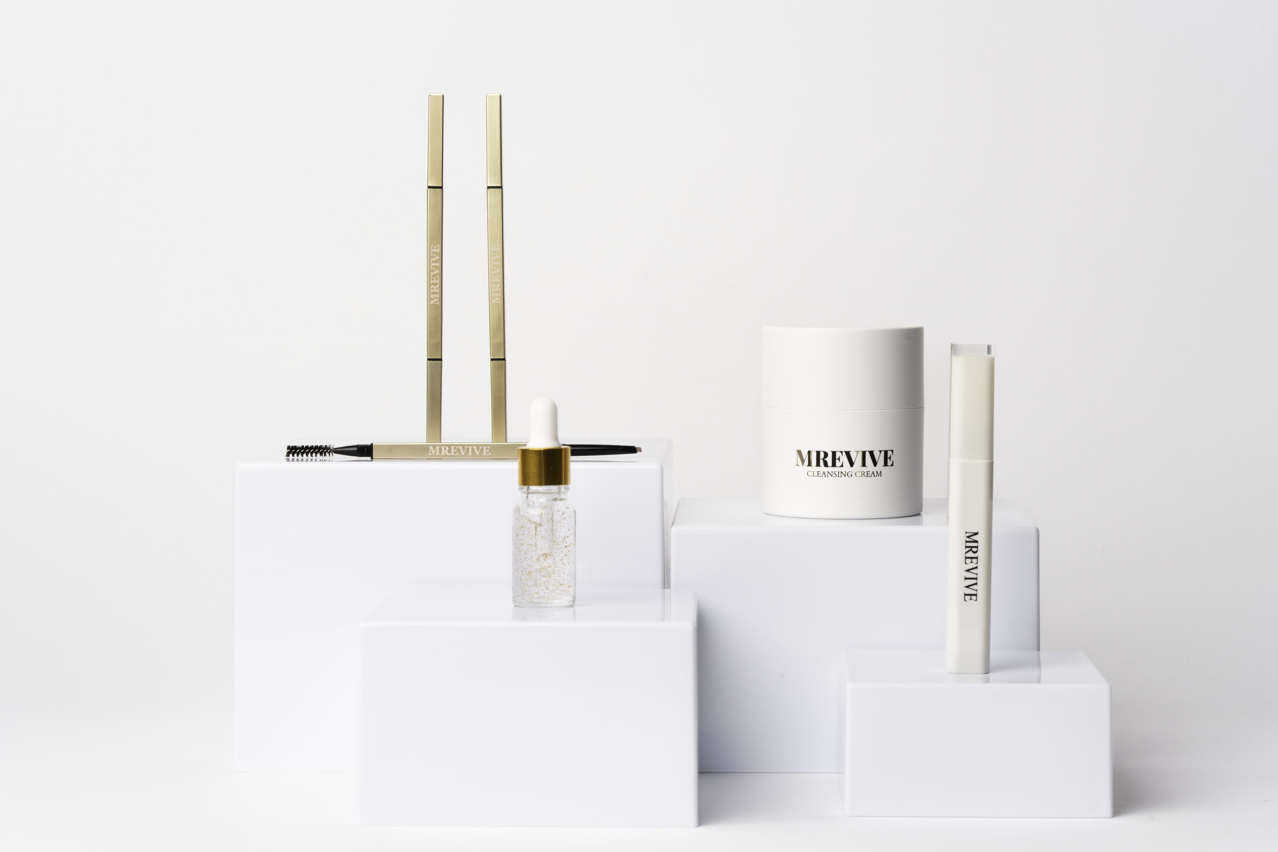 Skønhedsprodukter inklusive en flaske med perleformet serum, en hvid krukke med MREVIVE rensecreme, og to hvide rør med MREVIVE mærket, arrangeret på hvide platforme mod en hvid baggrund.