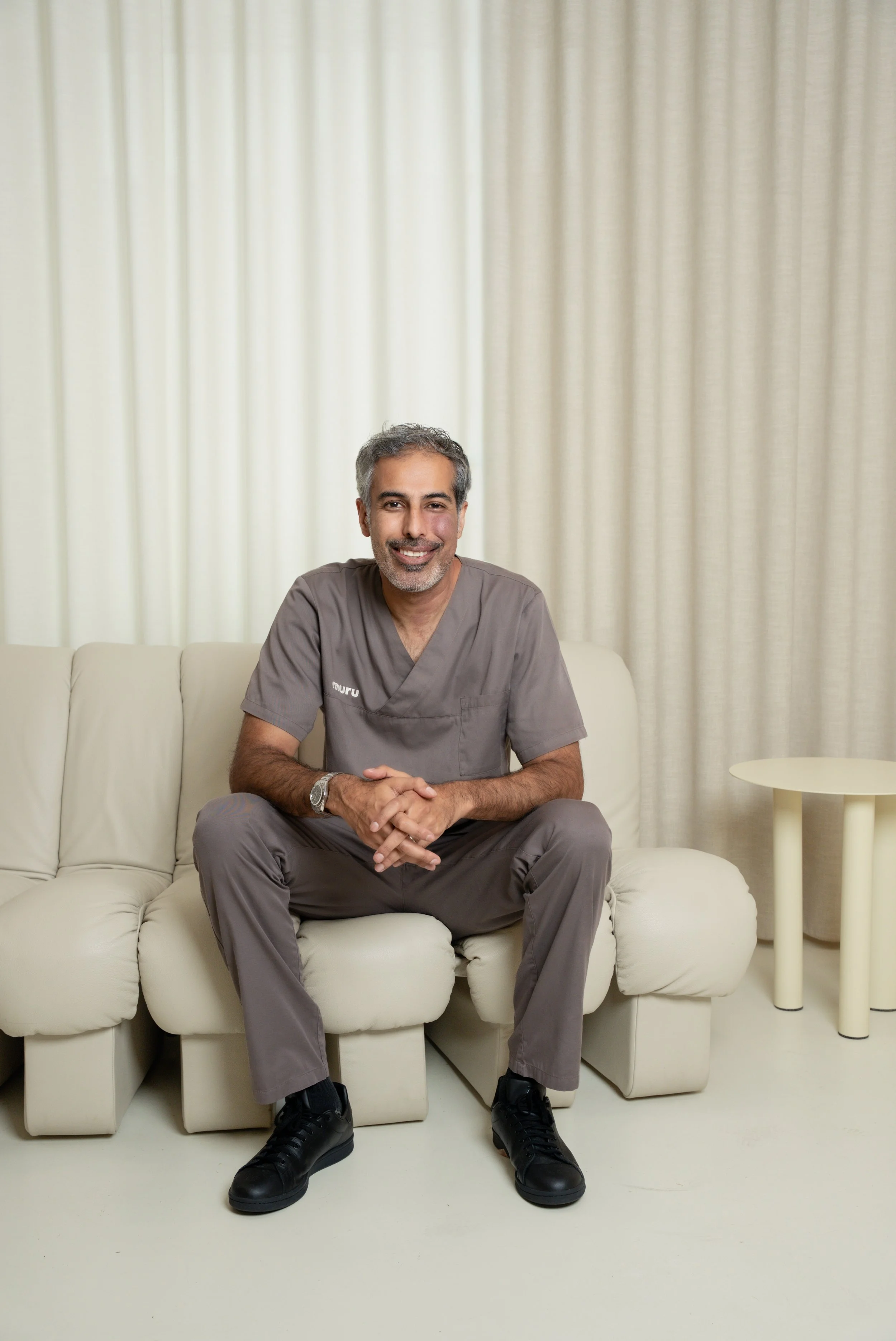 En mand i grå medicinsk uniform sidder på en hvid sofa med lukkede hænder og smiler. Bag ham er beige gardiner og en rund sidebord.