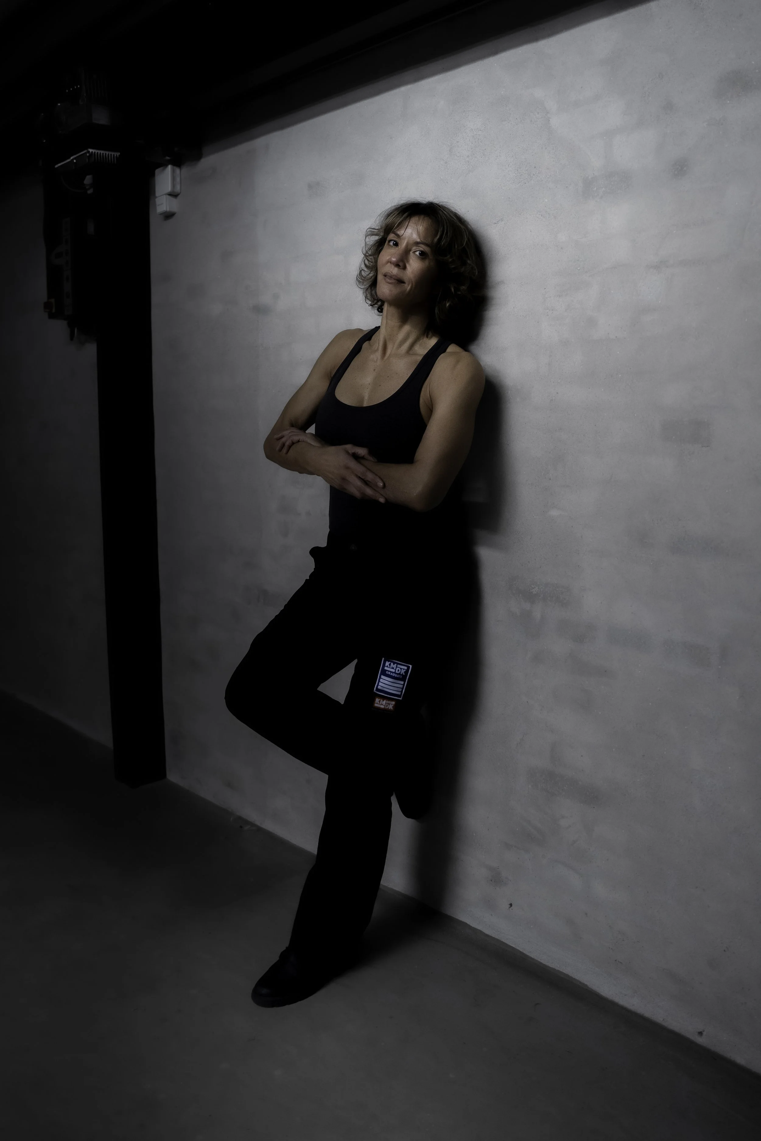 Kvinde i sort tøj står med armene over kors mod en hvid væg i et værelse med dæmpet belysning.
