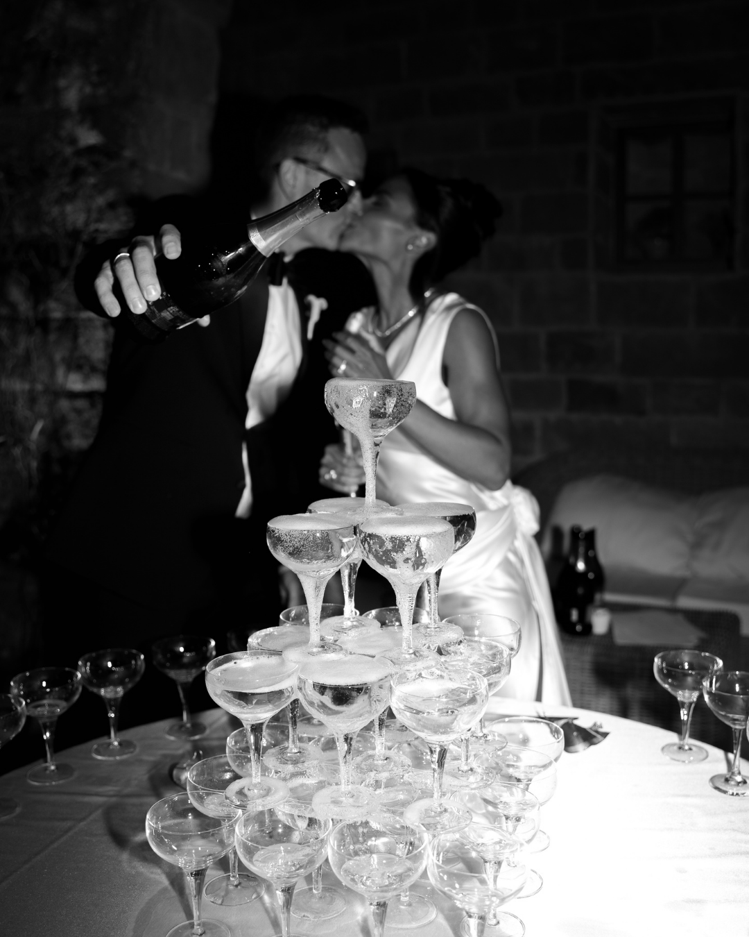 Et bryllupsbillede af et par, der kysser, med champagne i hånd, bag et tårn af champagneglas.