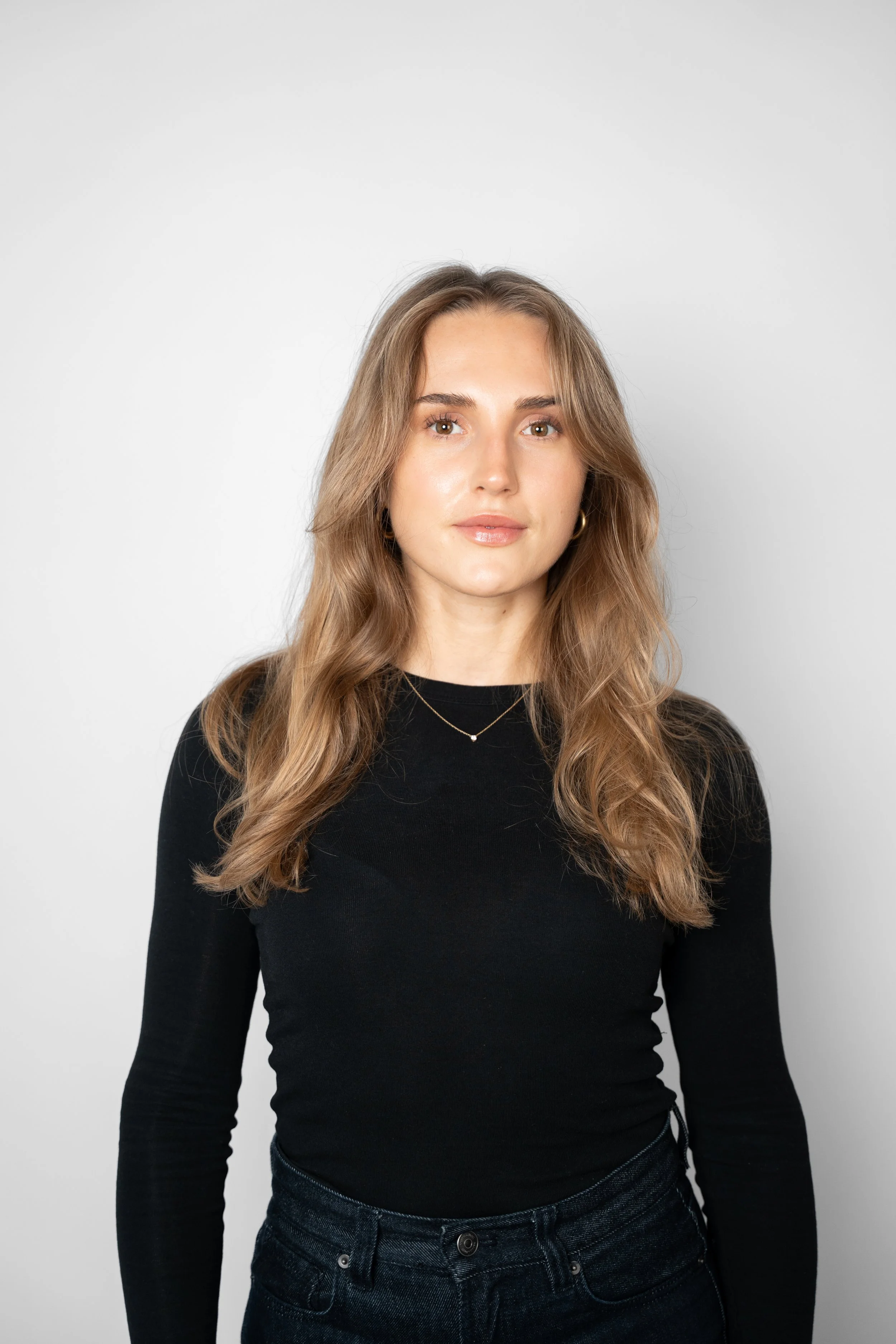 Et portræt af en ung kvinde med brunt, bølget hår, iført sort langærmet top og mørke jeans, stående foran en hvid baggrund.