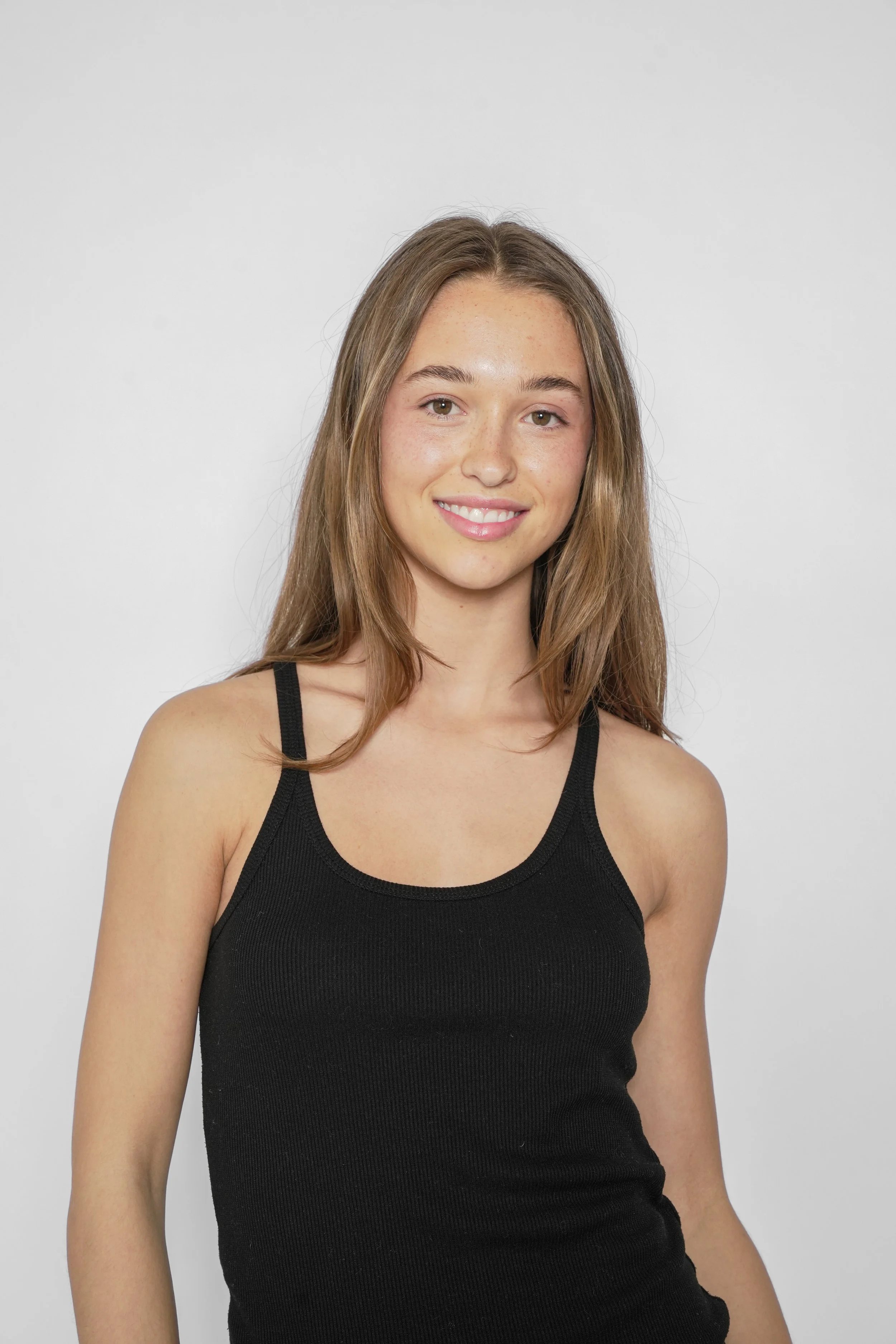 Et portræt af en smilende ung kvinde med langt brunt hår, iført en sort tanktop mod en enkel hvid baggrund.