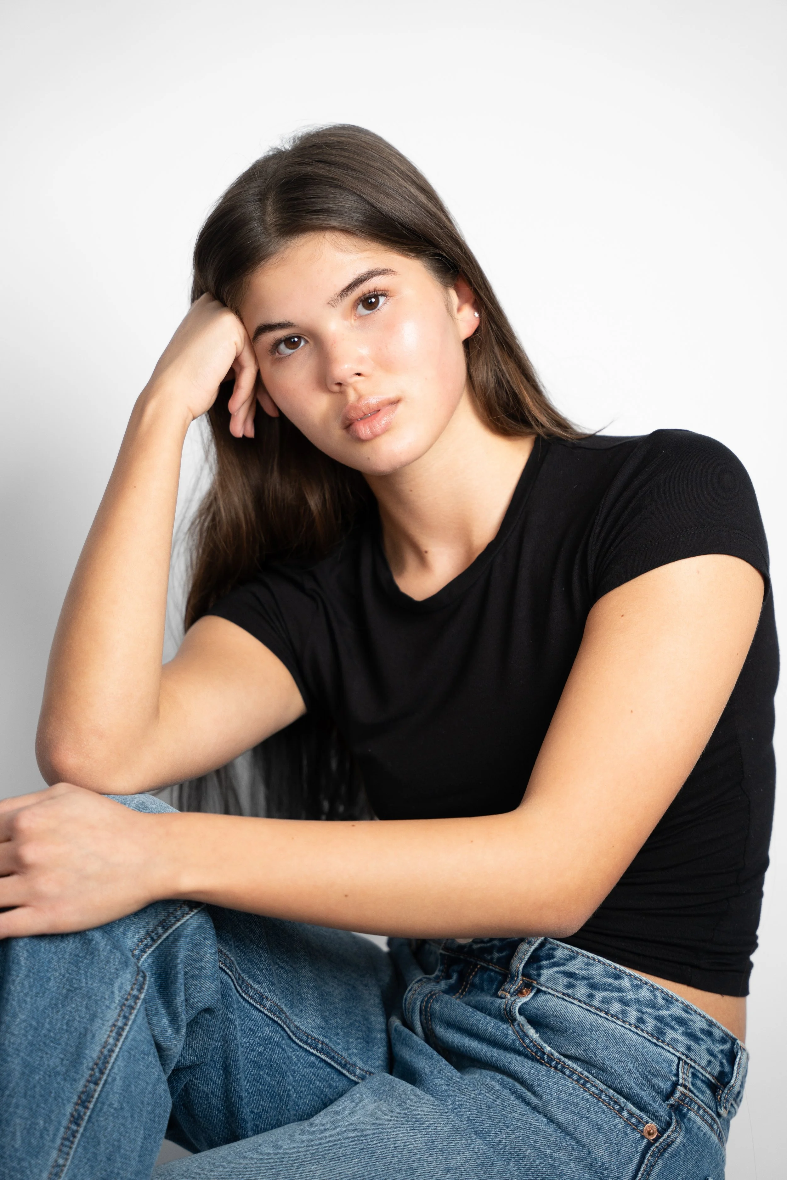 Ung kvinde med langt brunt hår, sort T-shirt og denim jeans sidder med arm hvilende på knæ, ser afslappet ud mod en hvid baggrund.