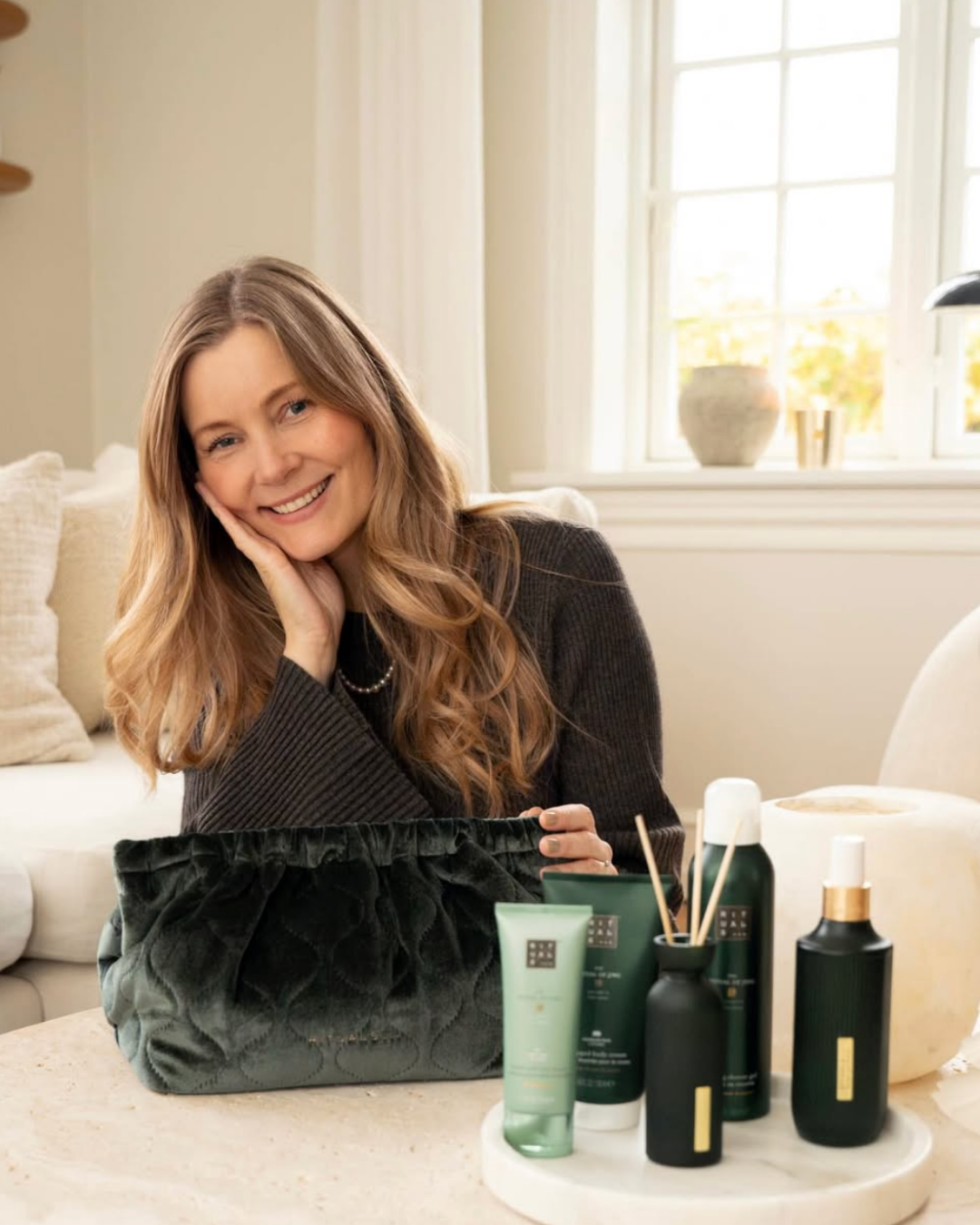 En smilende kvinde med langt, bølget, lysebrunt hår, sidder ved et bord med forskellige pleje- og skønhedsprodukter i sorte og grønne beholdere. Der er en stor, sort, quiltet taske foran hende og en hvid, rund bakke med flere beholdere. Bagved er et 