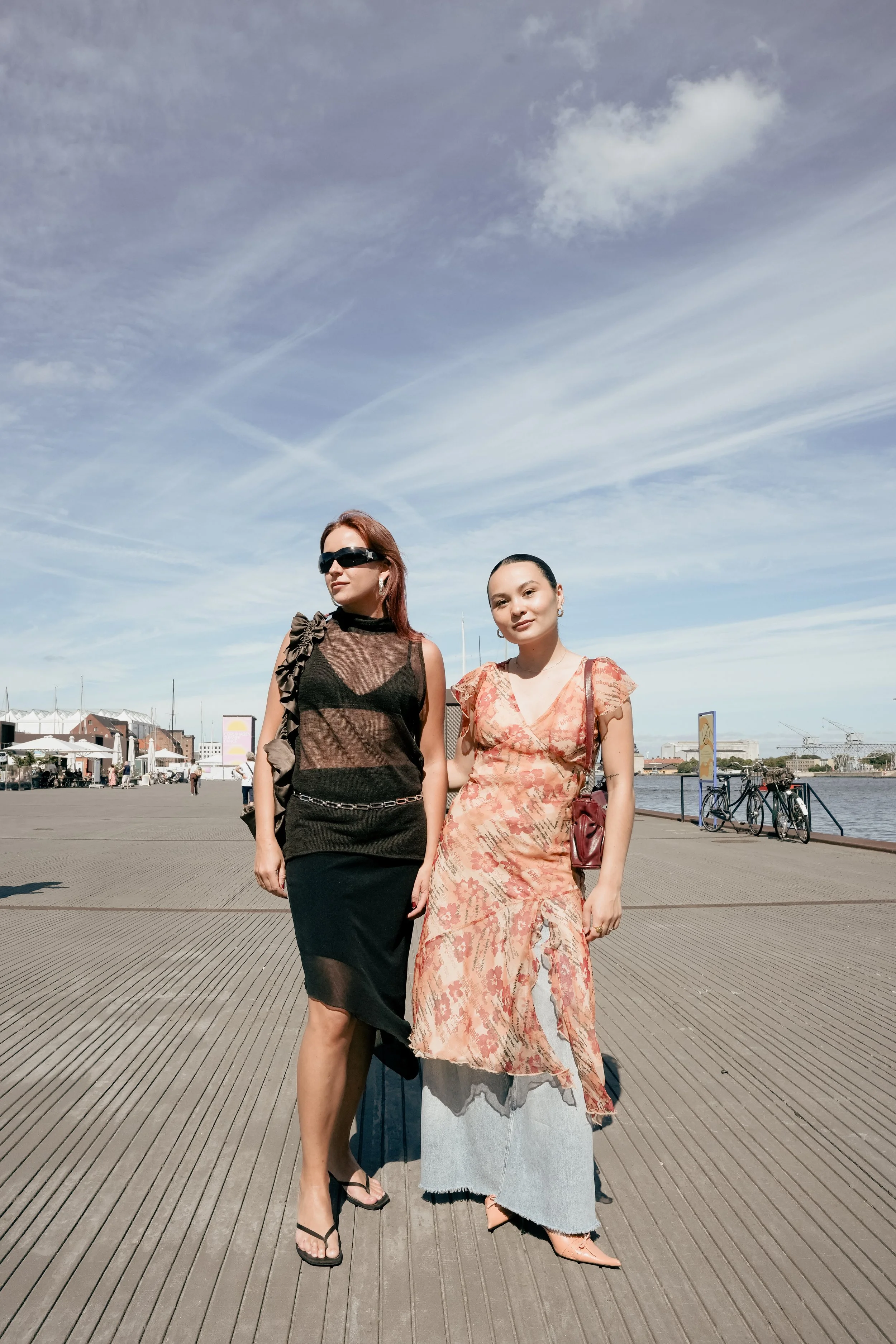 To kvinder går langs en havnefront på en solrig dag, klædt i fashionable tøj.