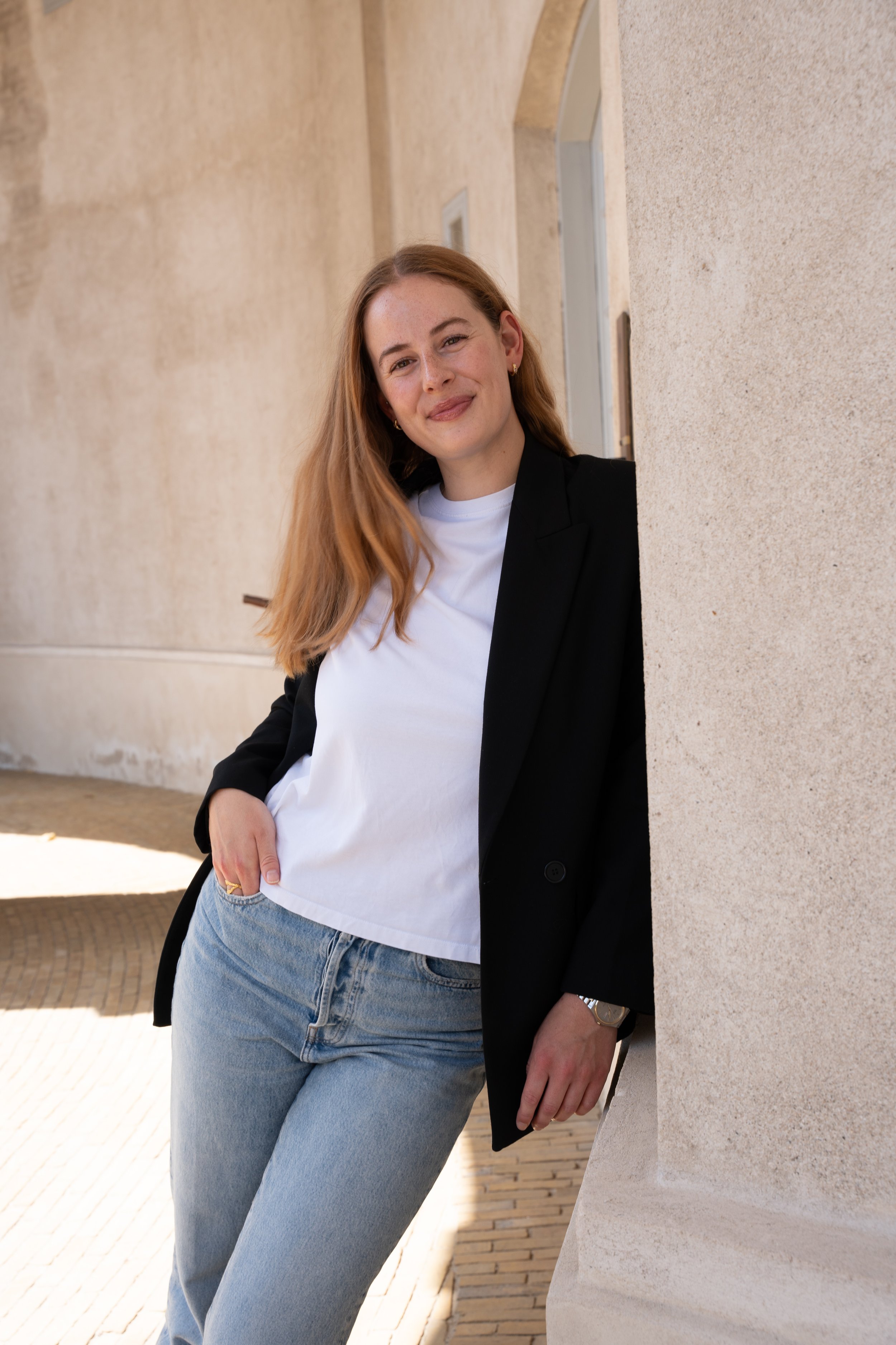 En kvinde med langt, lyst hår iført hvid T-shirt, sort blazer og jeans, står udendørs ved en væg og smiler.
