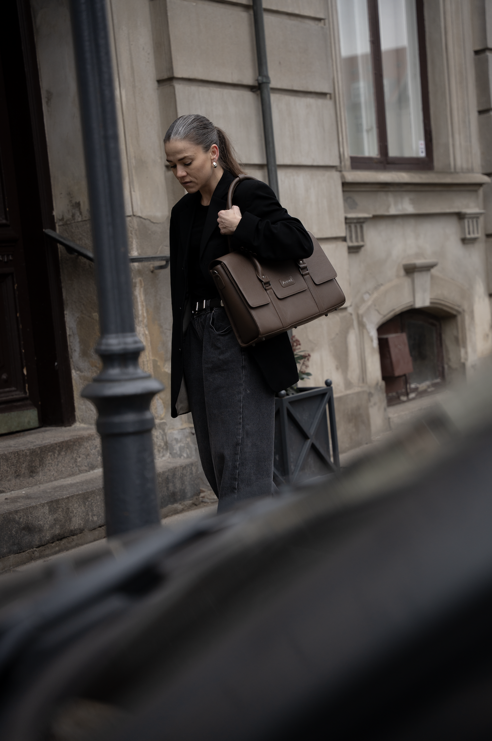 En kvinde med brunt hår bærer en stor brun taske og er iklædt en sort jakke og mørke bukser, står på en sten-trappe foran en gammel bygning i byen.