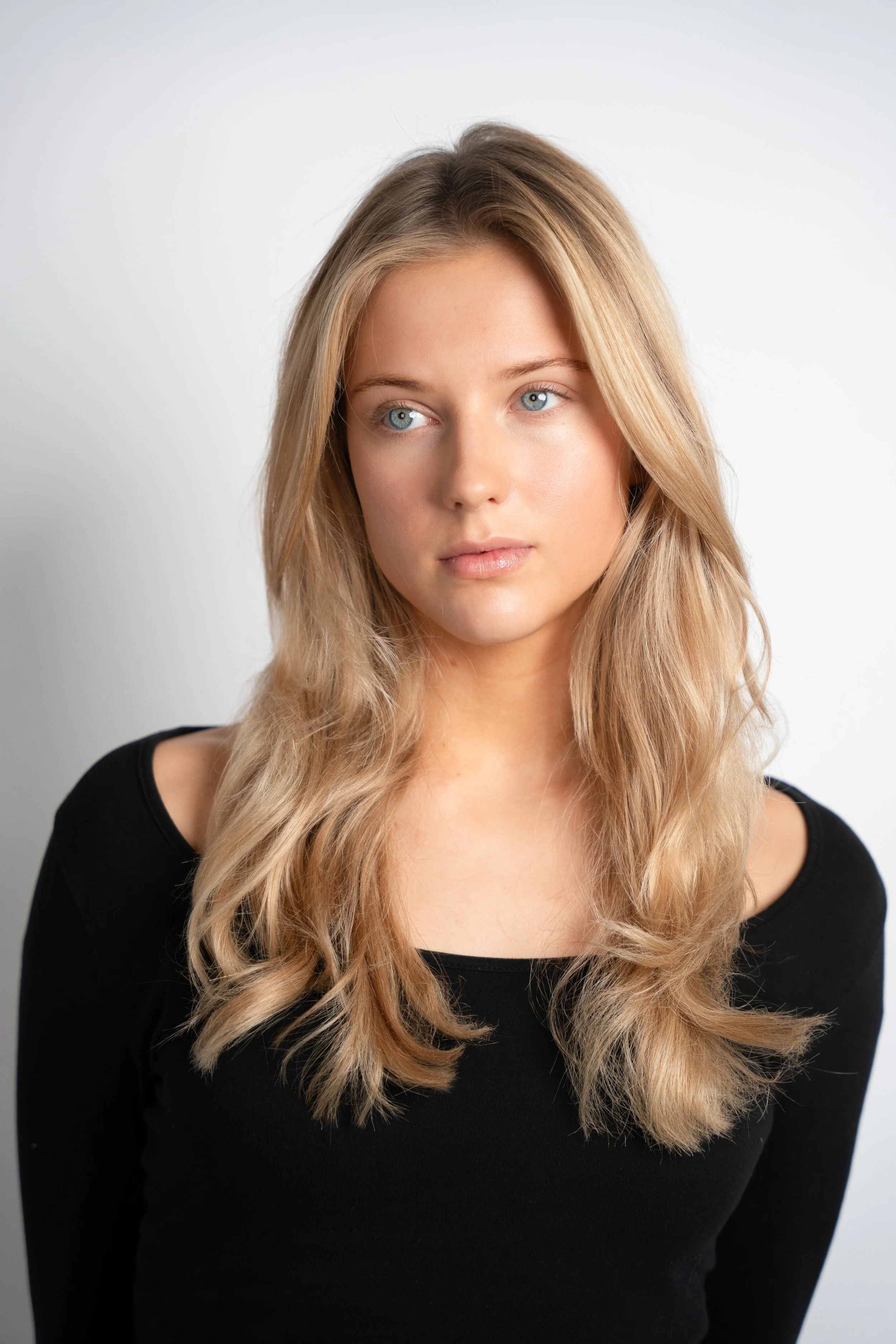 Portræt af en ung kvinde med lang, bølget blond hår iført en sort langærmet top, stående mod en hvid baggrund.