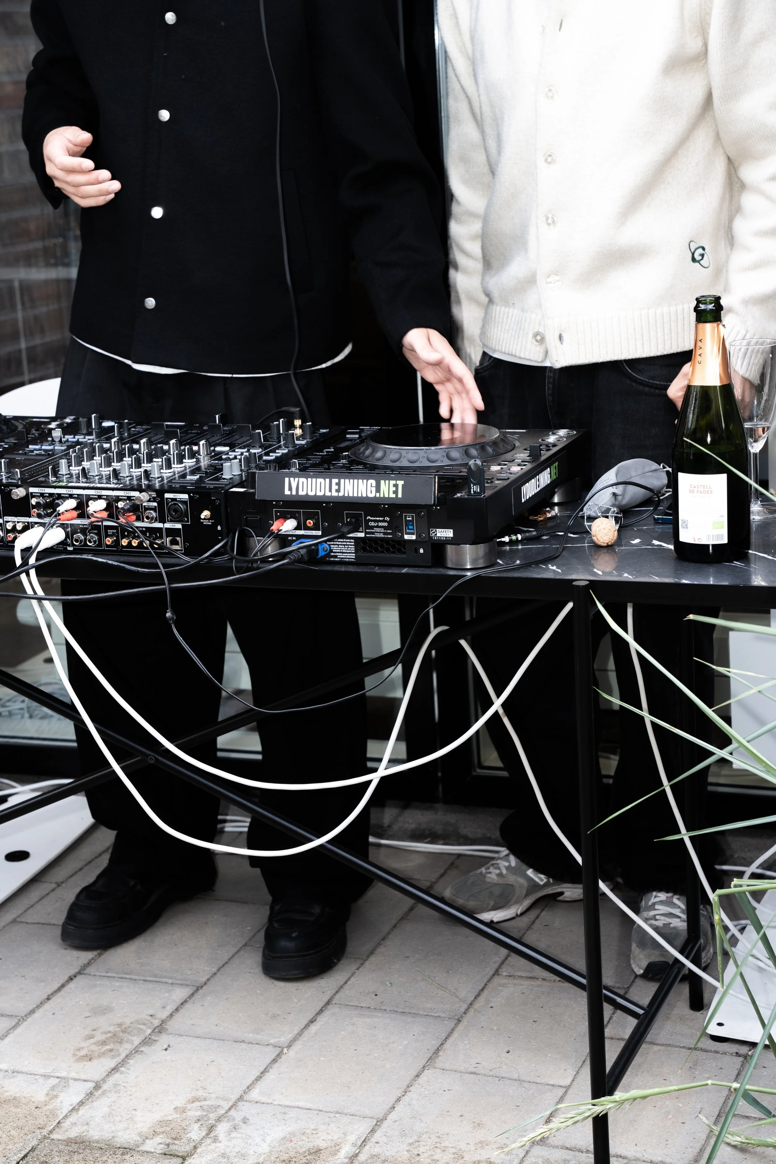 To personer, der står ved en DJ-opsætning med mixer og DJ-controller. Der er en flaske bobler og et glas på bordet.