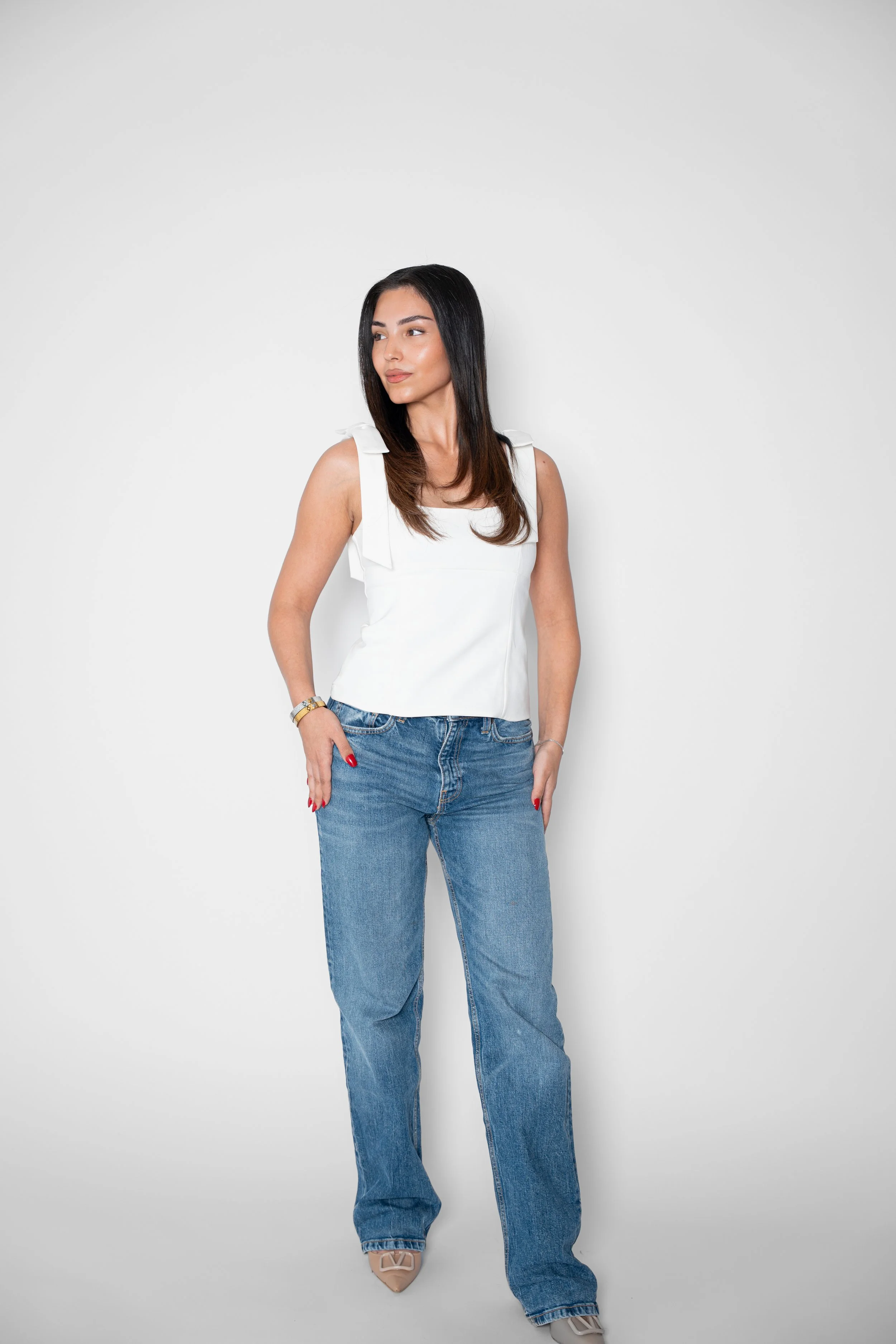 En kvinde står mod en hvid væg, iført en hvid top og blå jeans.