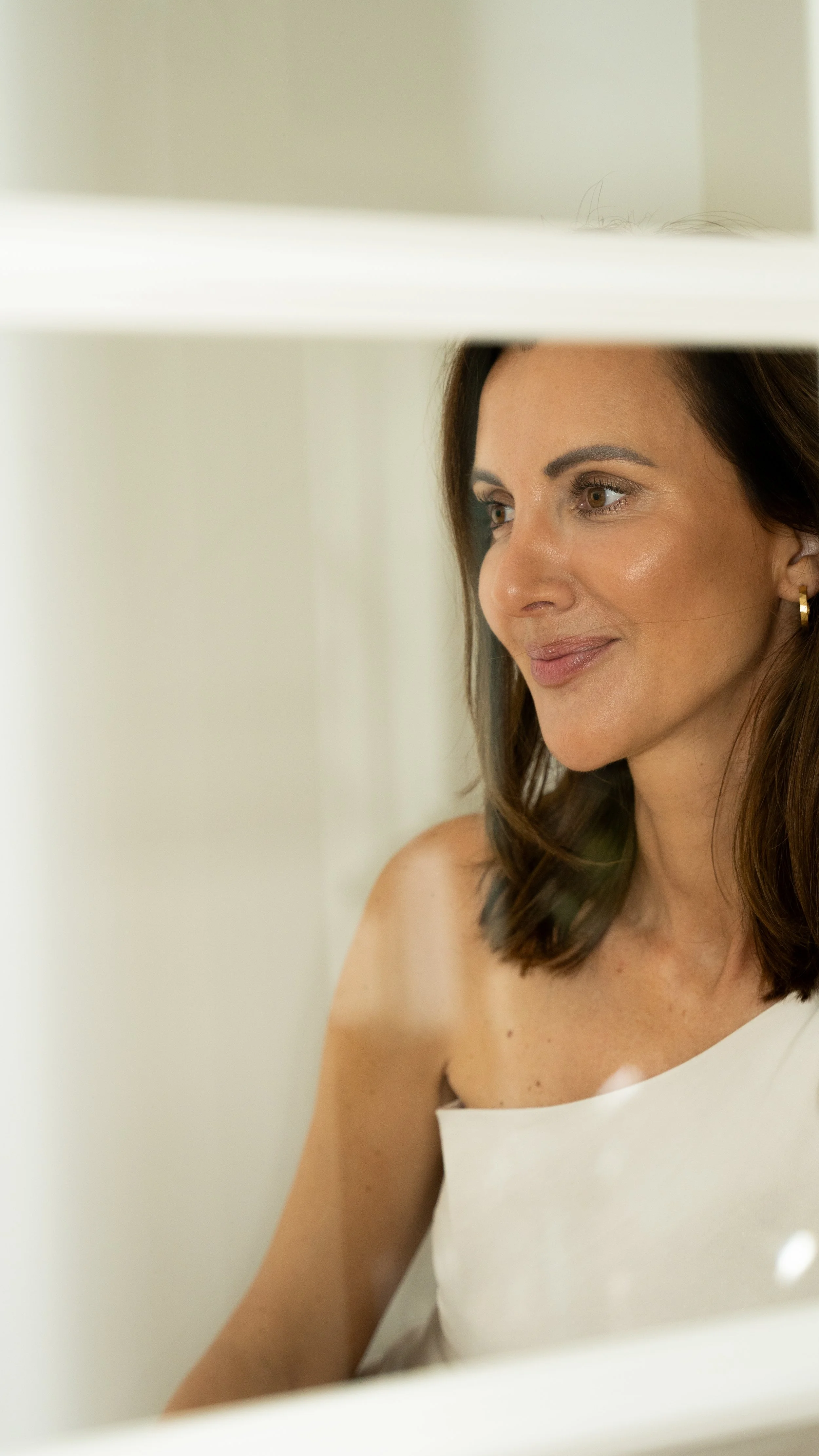 En kvinde med brunt, skulderlangt hår smiler blidt og ser ud gennem et vindue