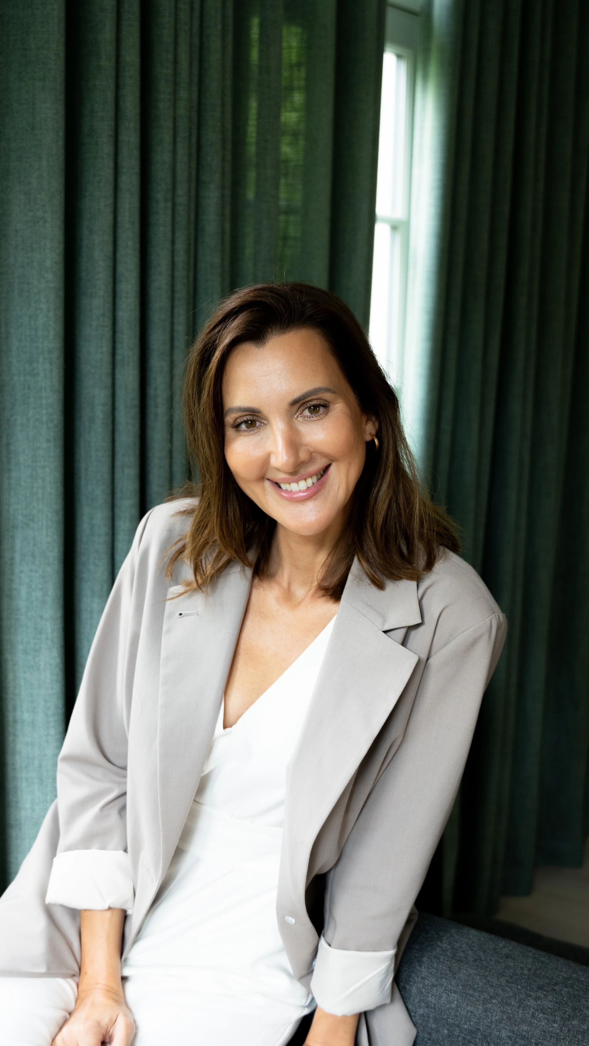 En smilende kvinde med mørkebrunt hår, iført hvid kjole og beige blazer, sidder foran grønne gardiner, op mod et vindue.
