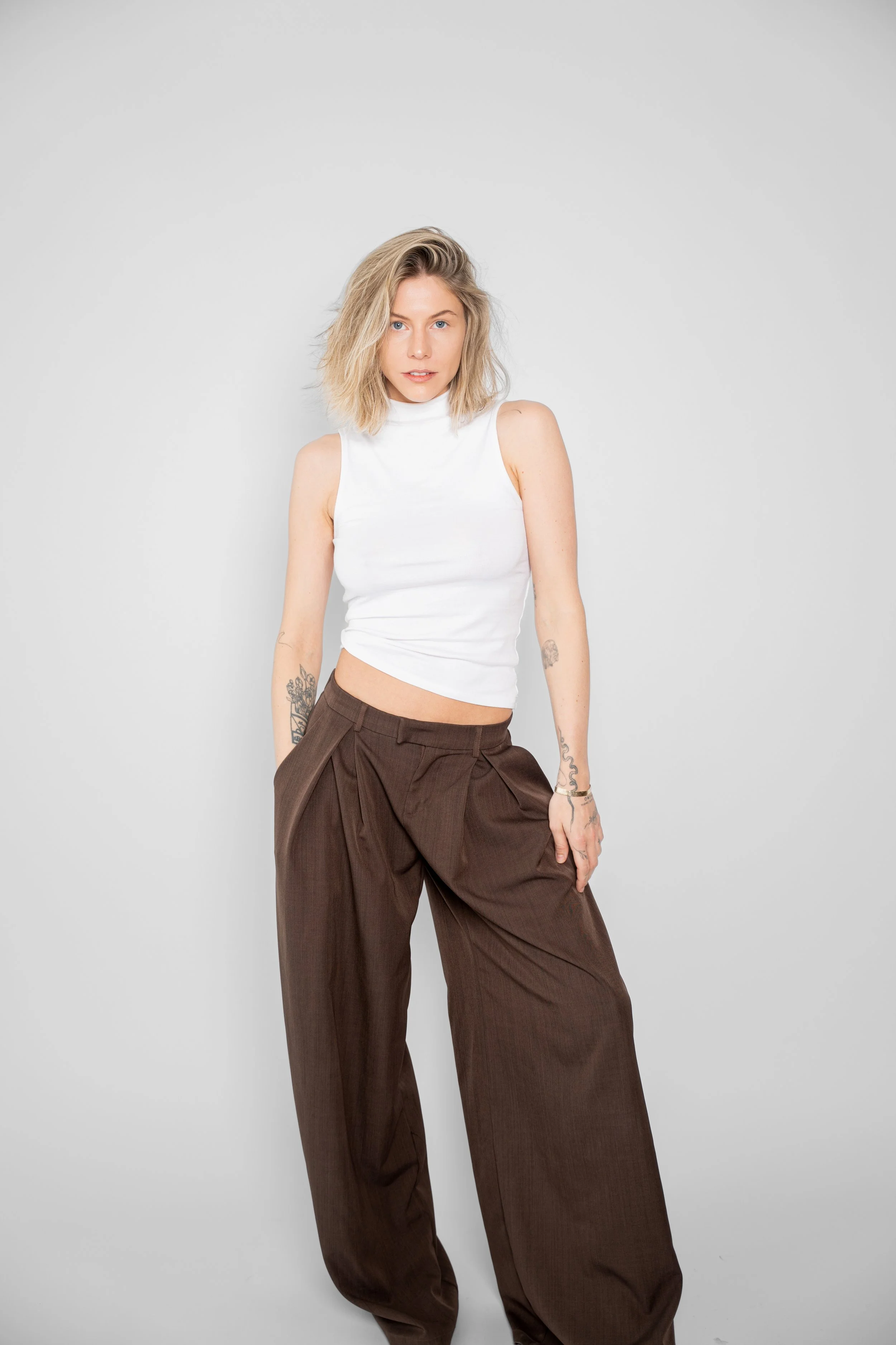 En ung kvinde med blond, skulderlangt hår, iført en hvid ærmeløs top og brede brune bukser, står foran en neutral grå baggrund.
