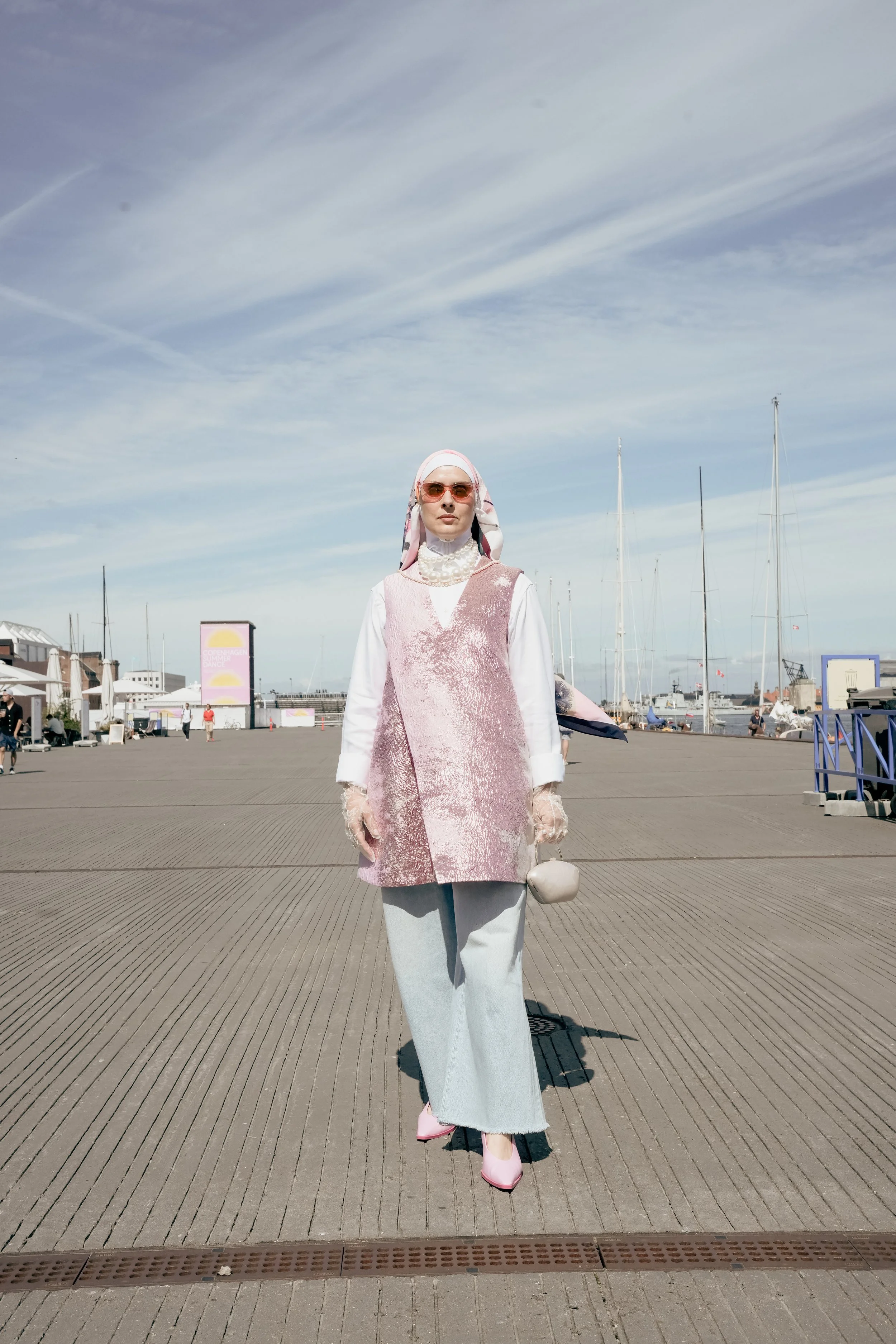 Kvinde i pastelfarvet outfit med solbriller står på en kaj ved vandet med lystbåde i baggrunden.