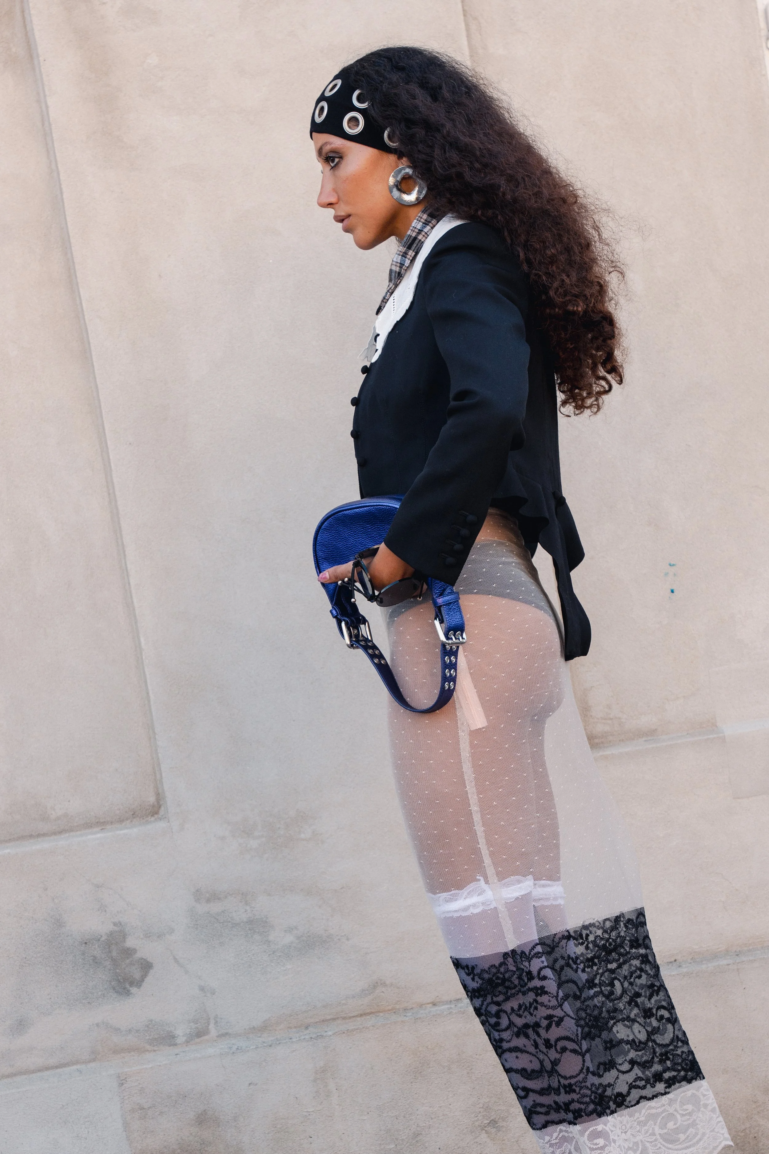 Kvinde med mørk krøllet hår iført sort bandana, stor ørering, sort blazer, og gennemsigtig tyl kjole med sorte blonder, holder blå taske.