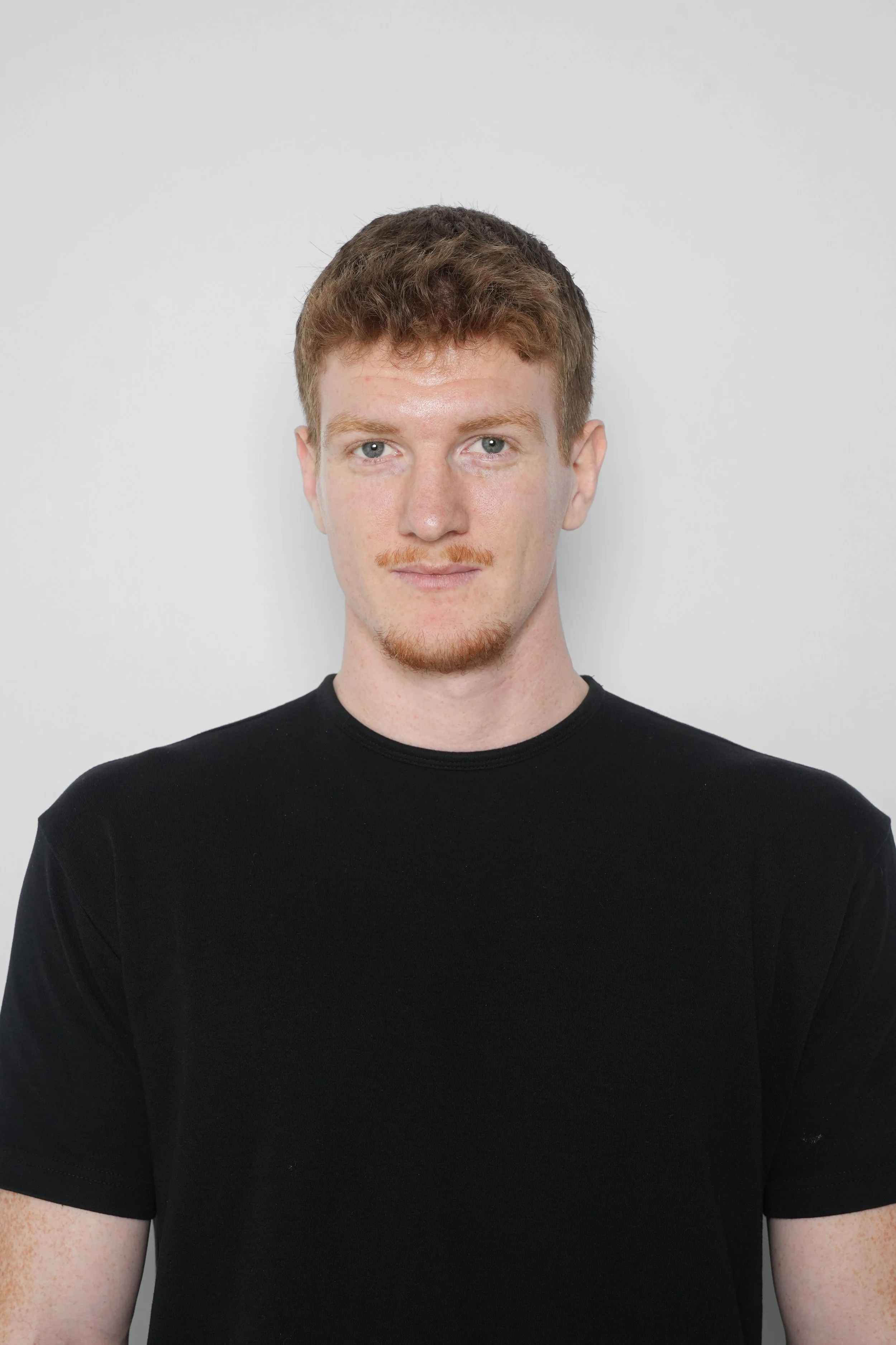 En ung mand med kort brunt hår og let skæg, iført sort t-shirt, står foran en hvid baggrund og ser direkte i kameraet.