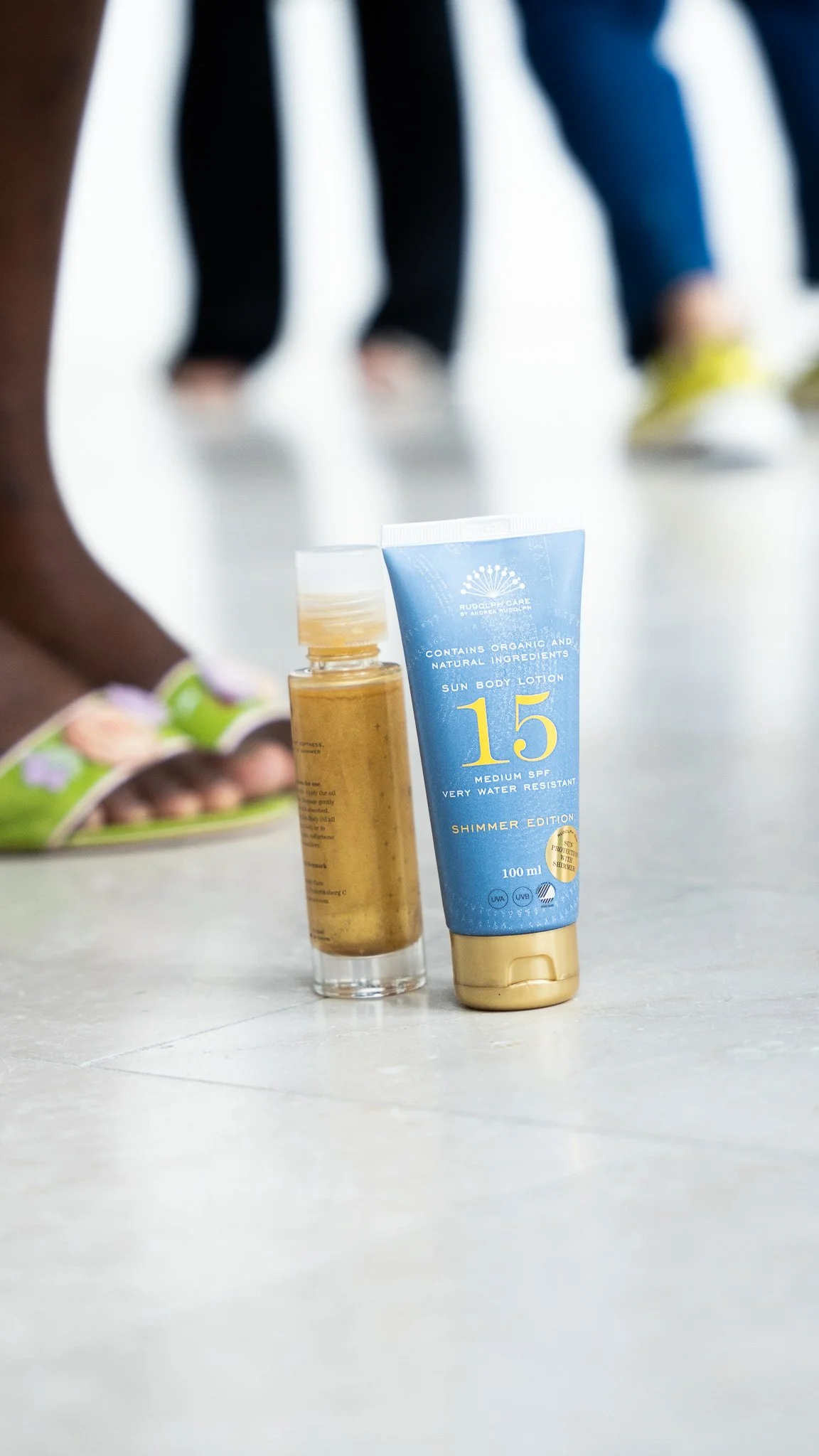 Solcreme og ansigtsolie placeret på gulvet med personer i baggrunden.