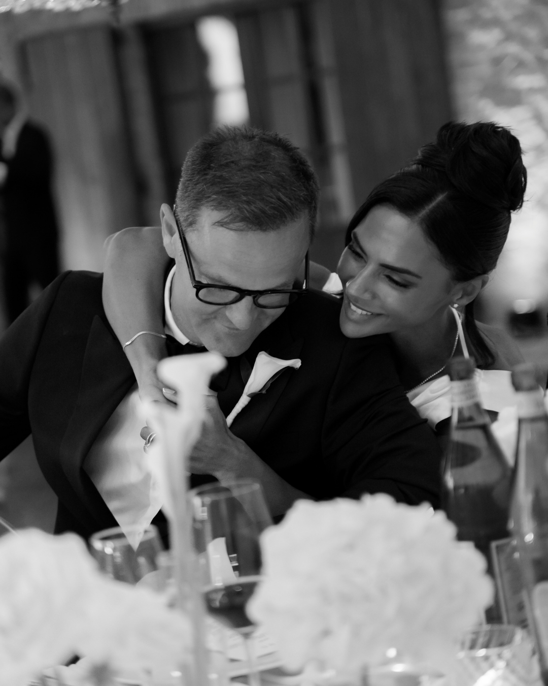 En sort-hvid foto af to personer, der sidder ved et bord ved en formel begivenhed, hvor den ene person er en mand med briller og tuxedo, og den anden person er en smilende kvinde med op-ordnet hår, der omfavner ham.