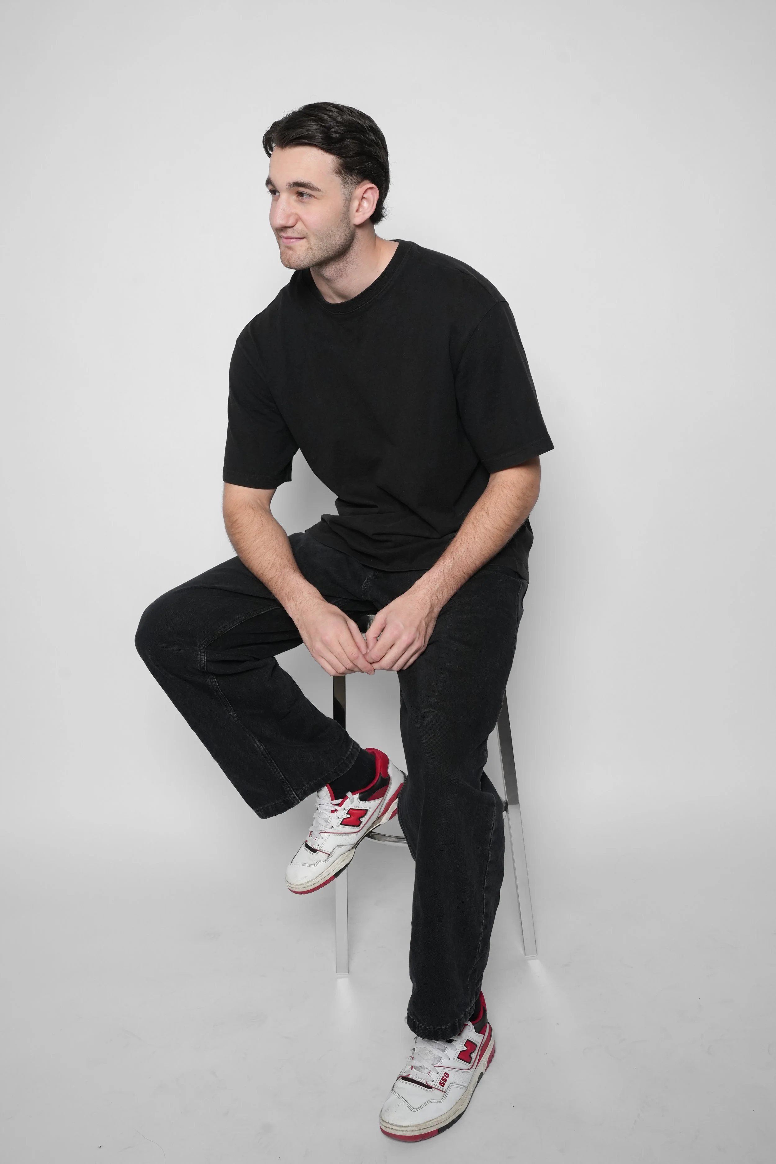 En mand sidder på en stol mod en hvid baggrund, iført sort t-shirt, sorte bukser og hvide sneakers med røde detaljer.