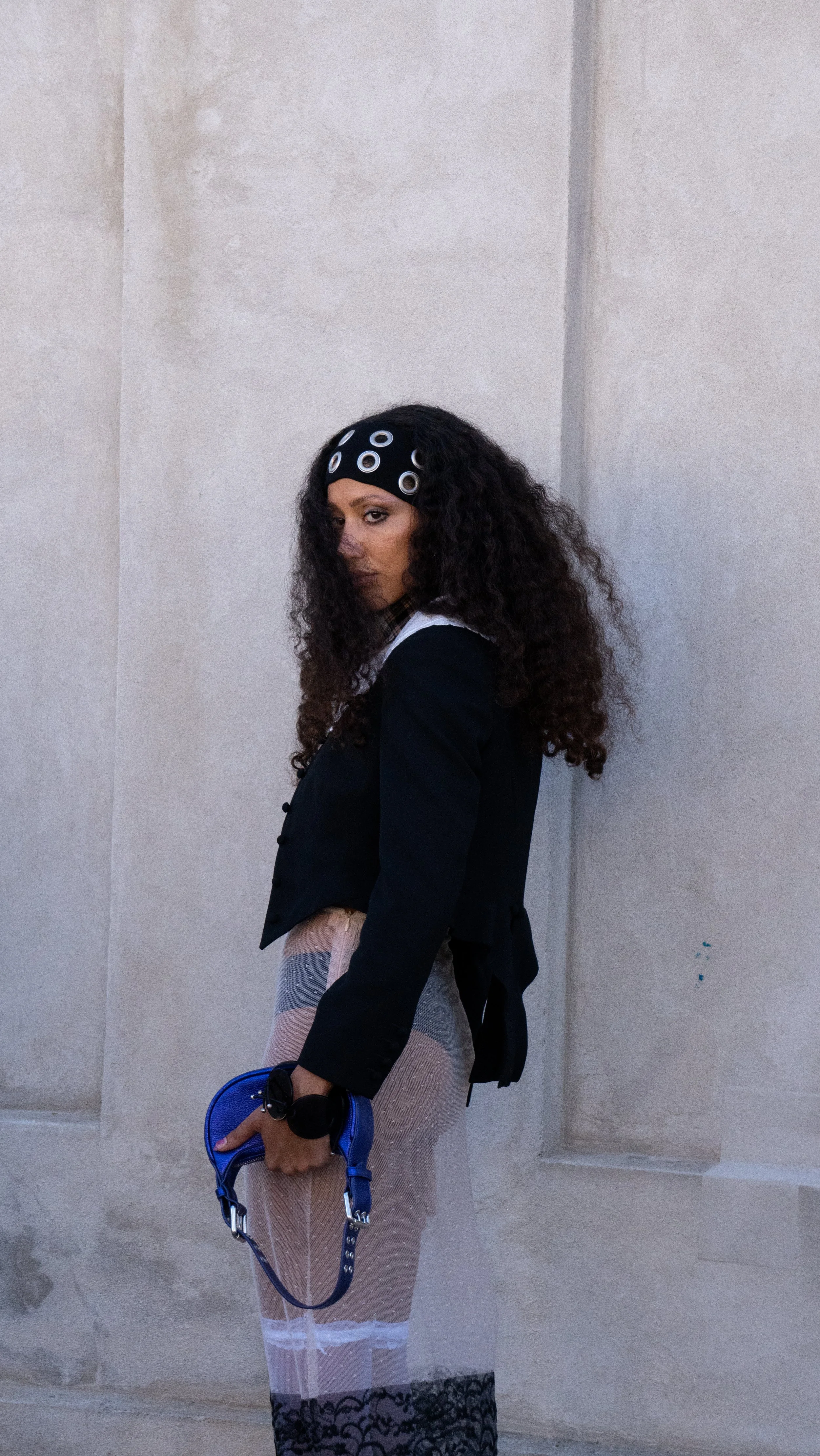 Kvinde med krøllet hår, mørk jakke og hvid kjole stående mod en murvæg.